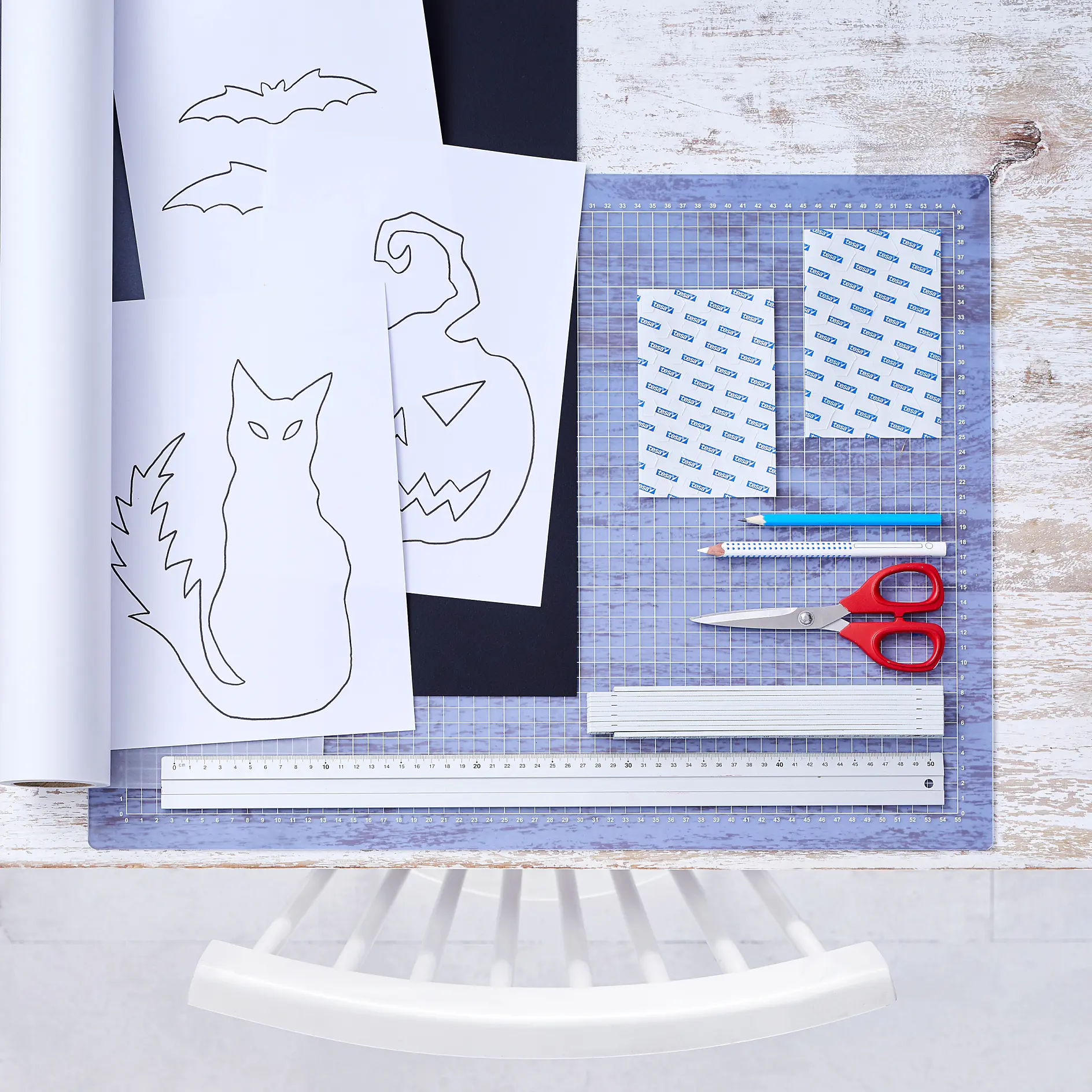 En arbetsyta med pysseltillbehör, inklusive en skärmatta, sax, linjal, blyertspenna, suddgummi, tesa-tejp och två hobbyknivar. Det finns svart och vitt papper med fladdermus-, katt- och pumpamotiv på. En stol syns i förgrunden. (This text has been generated by AI)