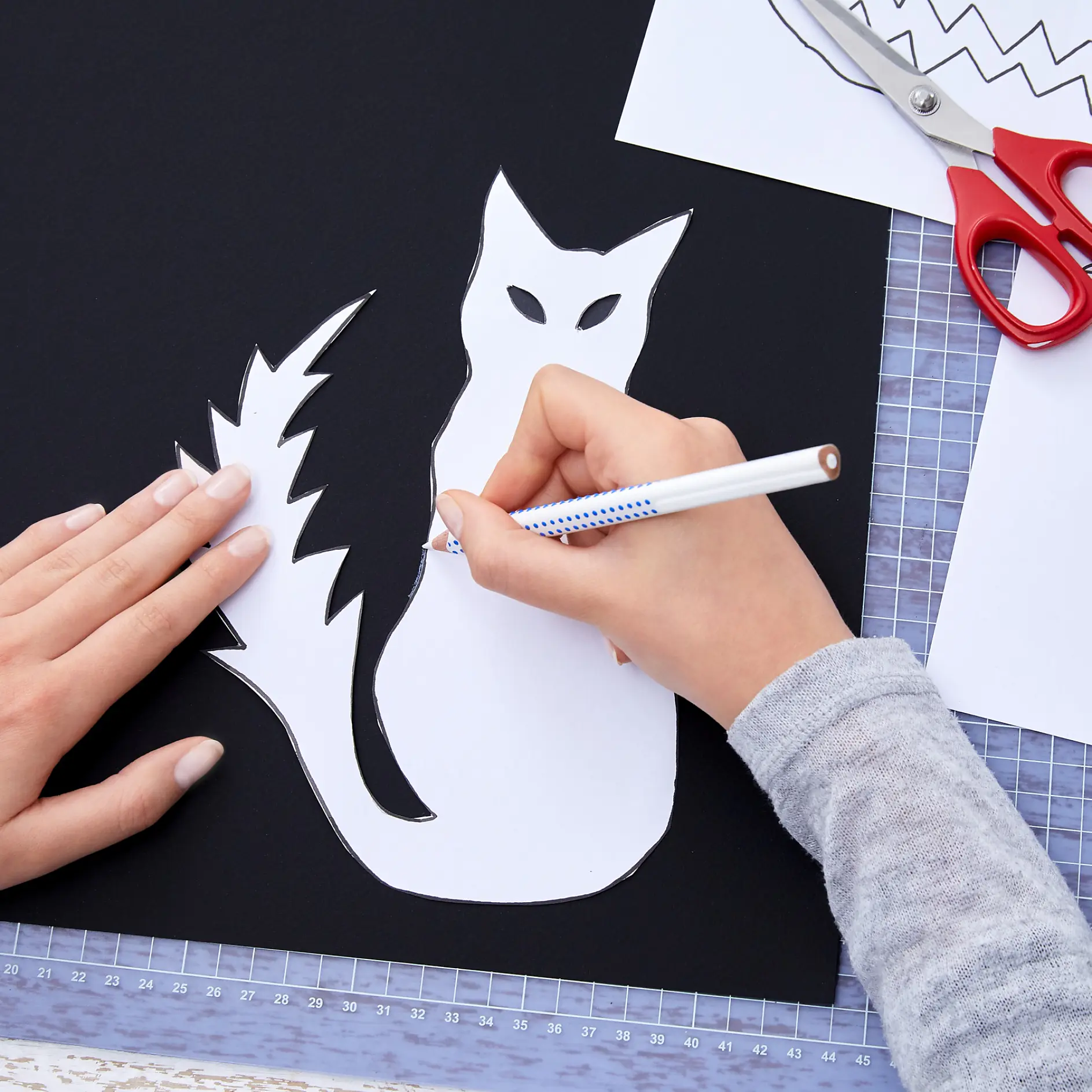 En person ritar av en kattsiluett på svart papper med en vit penna. Pappret ligger på en skärmatta, omgiven av saxar, tesa-tejp och en pappersmall. Personen bär en ljusgrå långärmad tröja. (This text has been generated by AI)