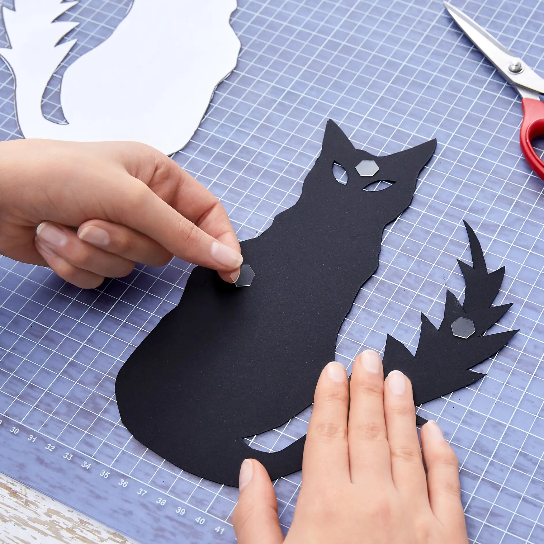 Händer sätter ihop svarta pappersutklipp av en sittande katt med yvig svans på en blå rutmönstrad matta. Vitt papper och röda saxar syns i bakgrunden tillsammans med rullar av tesa-tejp för pysselprocessen. Scenen visar hantverk eller DIY-konst. (This text has been generated by AI)