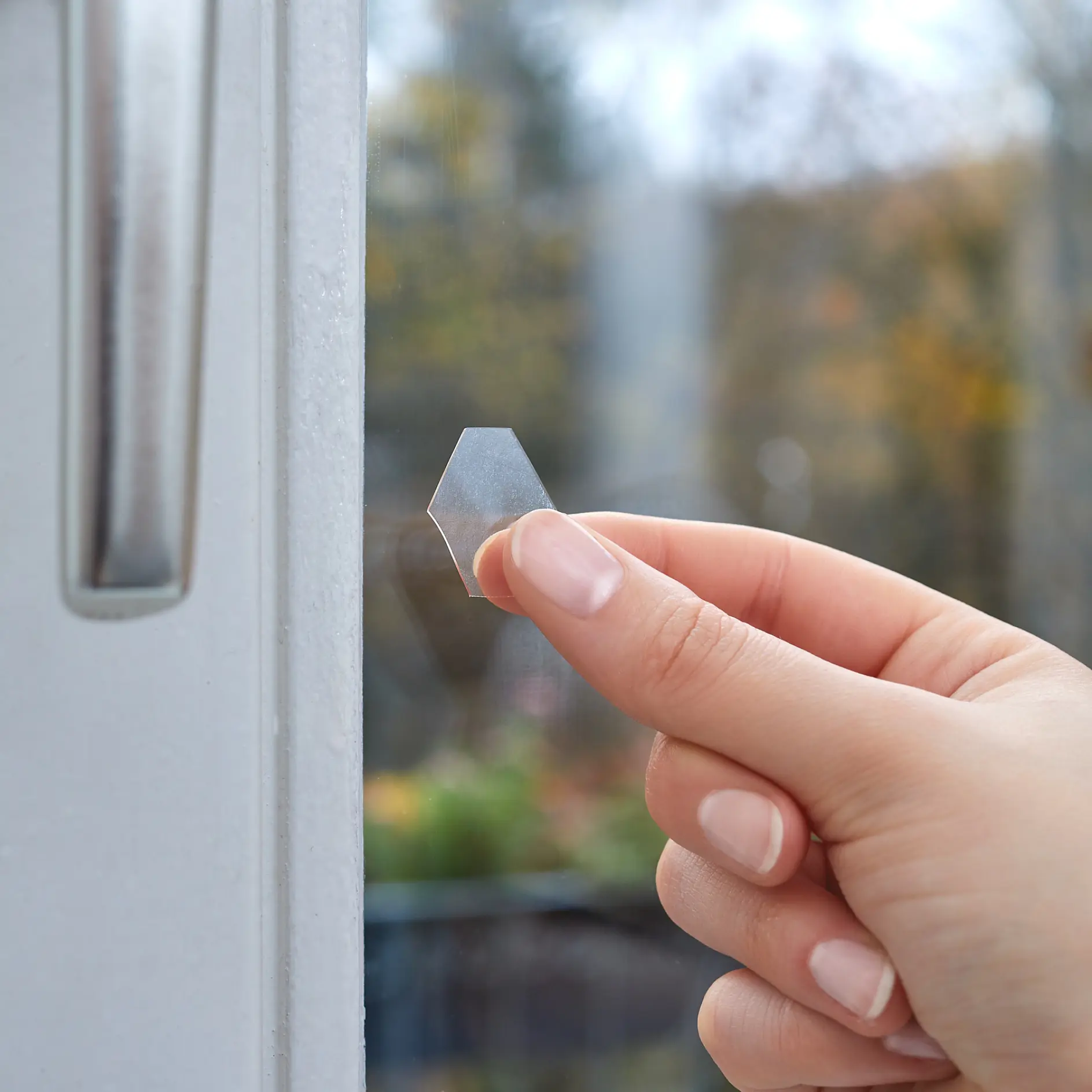 En hand håller en genomskinlig sexkantig tesa-tejp nära ett fönster. I bakgrunden syns en suddig utomhusscen med träd och grönska. (This text has been generated by AI)