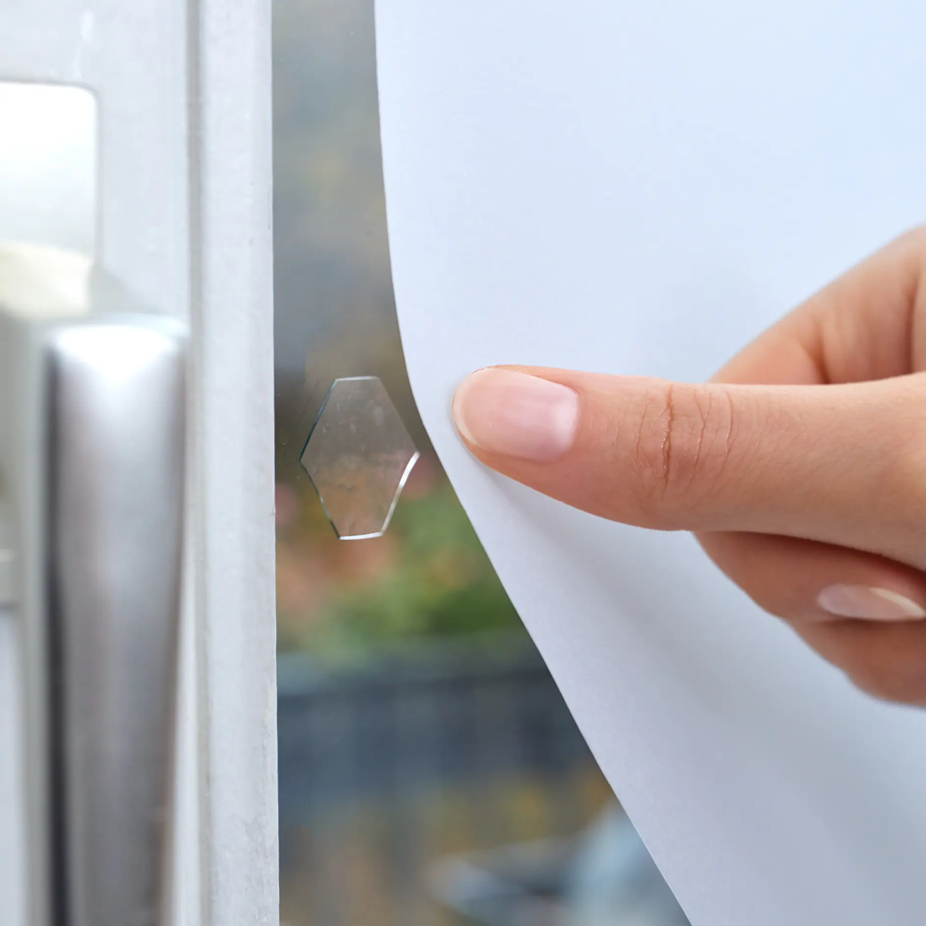 En hand trycker ett vitt pappersark mot en glasdörr. Bakom pappret syns en liten hexagonal bit tesa-tejp på glaset. Det silverfärgade dörrhandtaget är delvis synligt till vänster. Bakgrunden utanför dörren är suddig. (This text has been generated by AI)