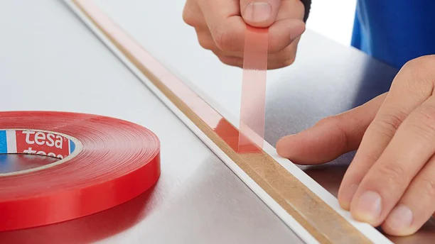 A person is applying a strip of red double-sided adhesive tesa tape to a flat surface. The tesa tape roll, labeled with tesa, is placed beside them. The persons hand is pulling the tesa tape to stick it down firmly. The surface appears metallic and smooth. (This text has been generated by AI)