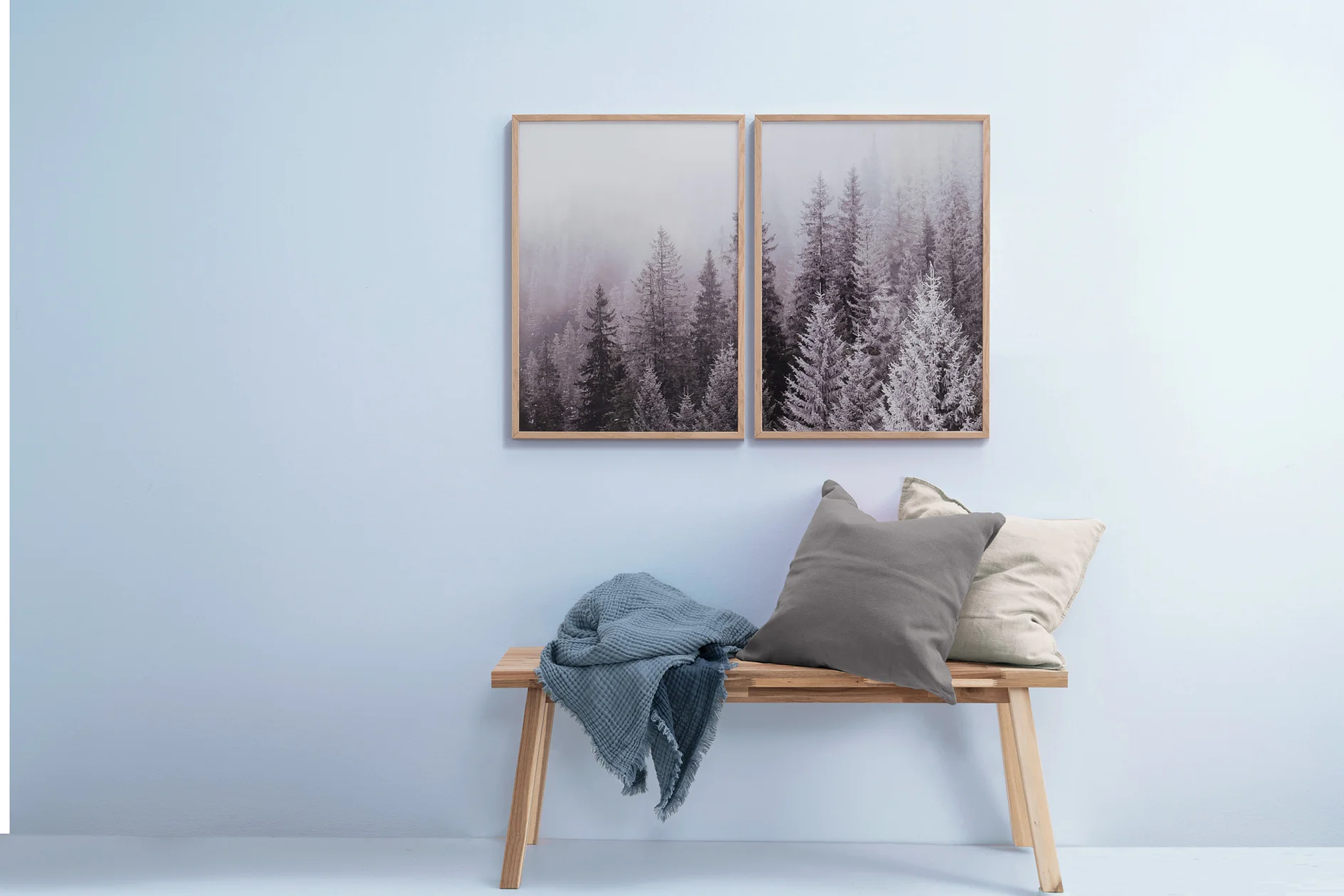 En träbänk med en grå och beige kudde och en blå slängfilt står mot en ljusblå vägg. Två inramade bilder av frostade vintergröna träd hänger ovanför bänken, var och en fäst med tesa-tejp. (This text has been generated by AI)