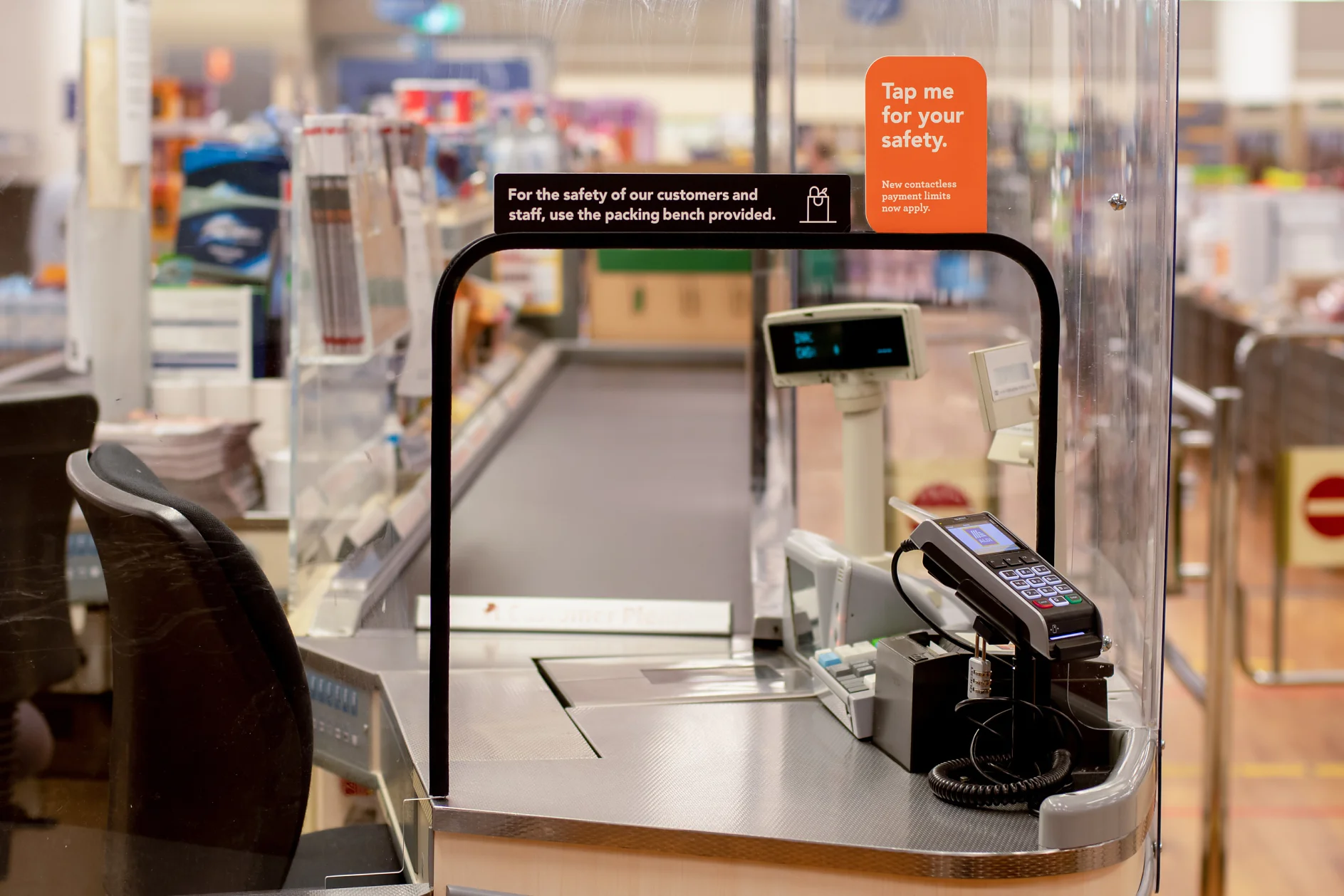 Självutcheckningsstation i en stormarknad med plexiglasbarriär. En digital skärm och en kortbetalningsterminal syns. På en orange skylt står det tesa tejpa mig för din säkerhet. Nya sanitetsprotokoll har tillämpats. (This text has been generated by AI)