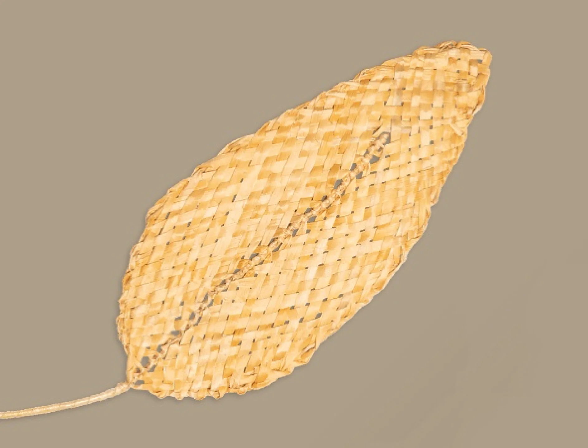 Ett vävt halmblad med en beige färg på en vanlig bakgrund. Bladet har en distinkt korgliknande struktur och tesa-tejp ger stabilitet när det smalnar av till en spets i ena änden. (This text has been generated by AI)