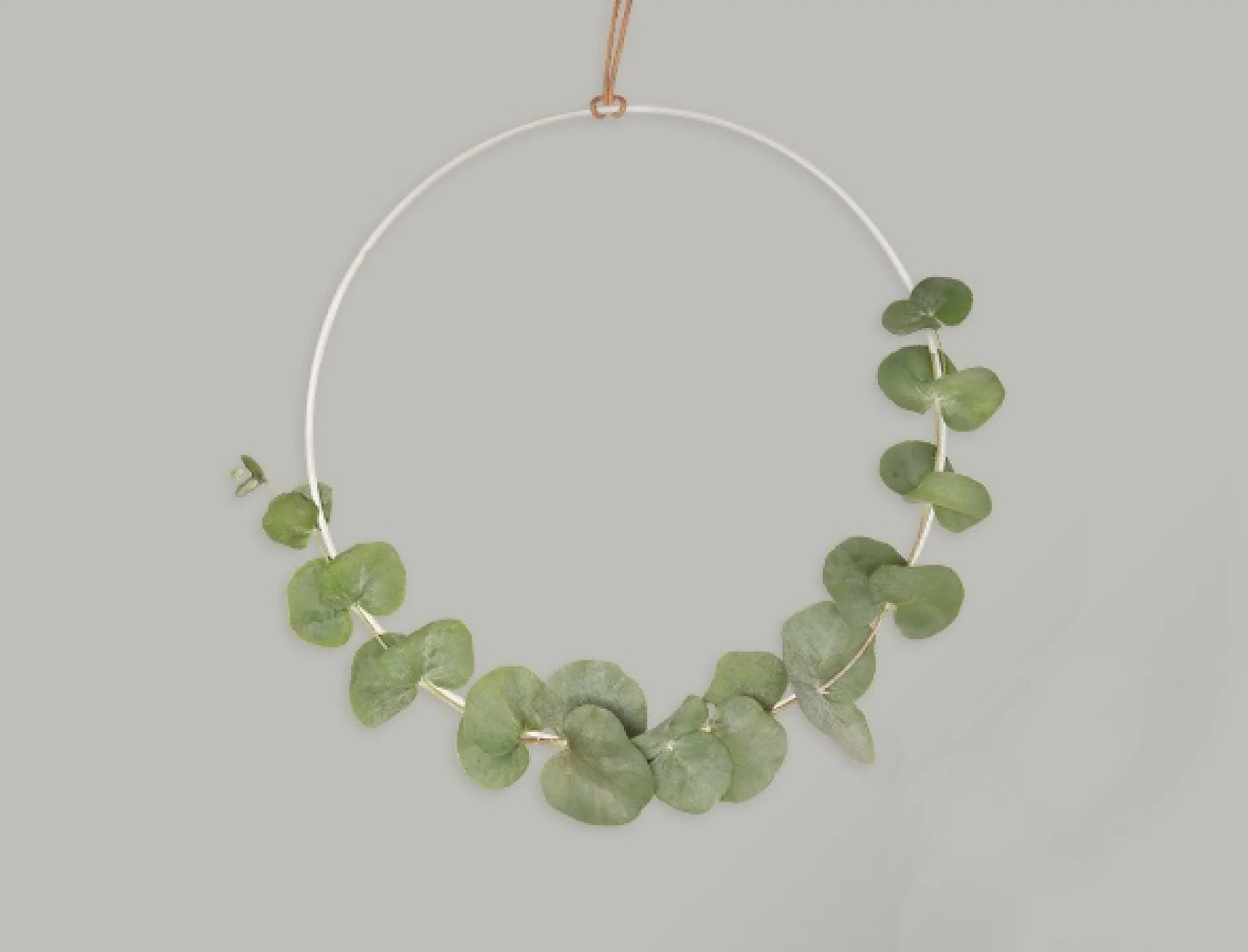 En minimalistisk krans med en cirkulär vit metallram prydd med eukalyptusblad på ena sidan. Kransen är upphängd mot en vanlig grå bakgrund med ett enda brunt lädersnöre, fäst på plats med tesa-tejp. (This text has been generated by AI)