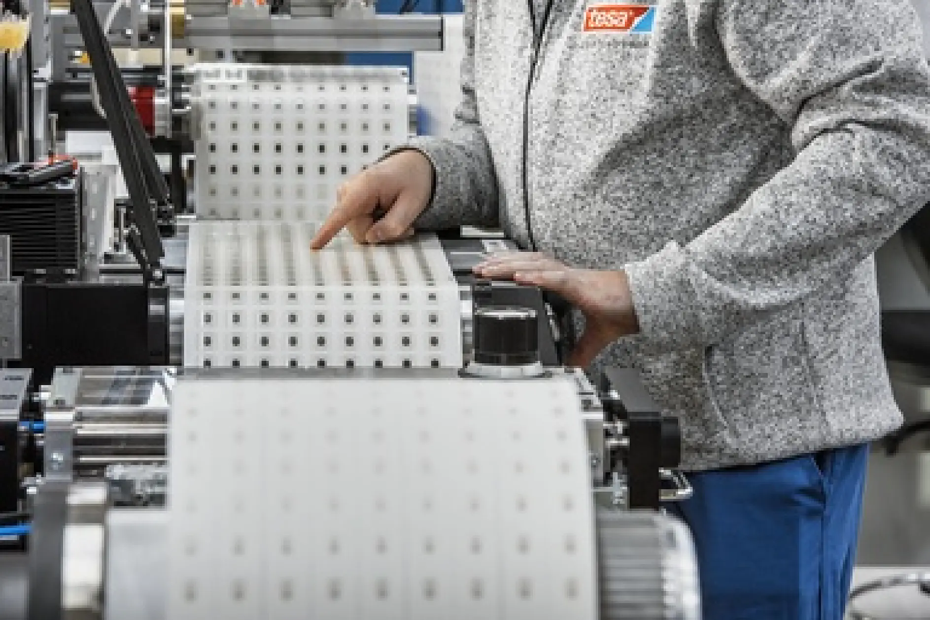 A person wearing a gray sweater operates a machine with large rolls of tesa tape, featuring square cutouts. The workspace appears industrial, with a focus on precision handling of the material. (This text has been generated by AI)