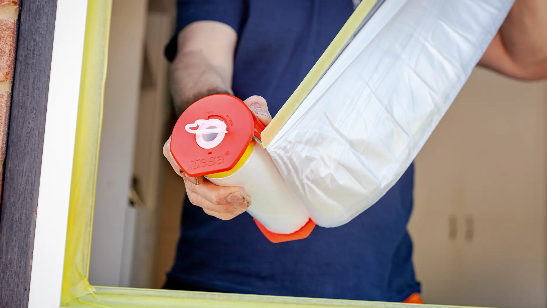 Person holding a roll of masking film with a red handle near a window frame. The window edges are partially covered with yellow tesa tape, presumably for painting or renovations. The person is wearing a dark blue shirt. (This text has been generated by AI)