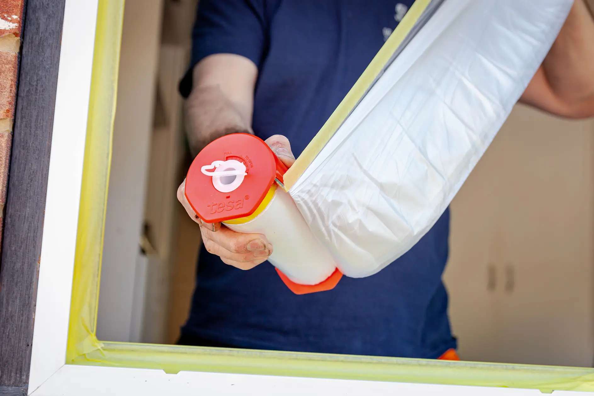 Person holding a roll of masking film with a red handle near a window frame. The window edges are partially covered with yellow tesa tape, presumably for painting or renovations. The person is wearing a dark blue shirt. (This text has been generated by AI)