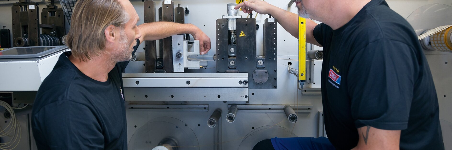 Two men are kneeling in front of industrial machinery, discussing its components. Both are wearing black shirts and blue pants. One man points at a part wrapped with tesa tape, while the other looks on attentively. The setting appears to be a workshop or factory. (This text has been generated by AI)