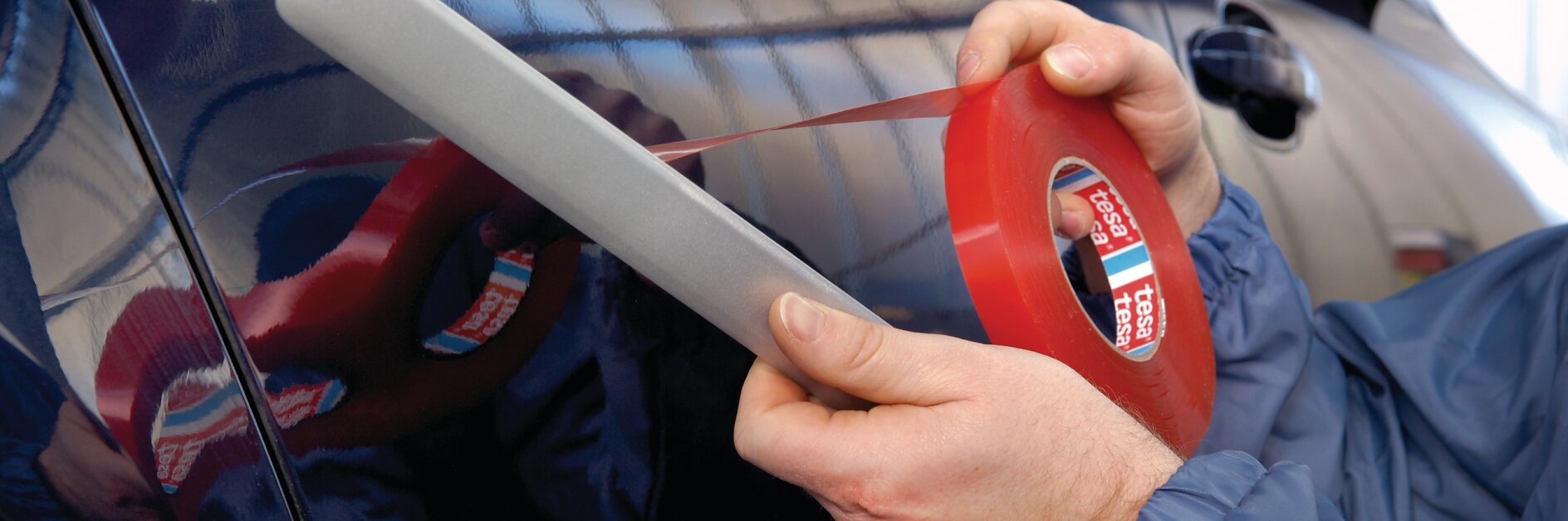 A person in a blue jacket is applying red tesa tape to the side of a dark-colored car. They are holding a tool with the tesa tape to ensure even application. The background shows a blurry interior of a garage or workshop. (This text has been generated by AI)