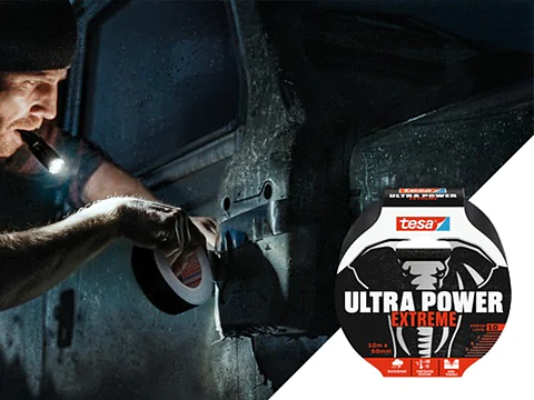 A person uses a flashlight to apply black tesa tape to a vehicle door in a dimly lit setting. The tesa tape is shown in the foreground with the label tesa Ultra Power Extreme featuring an elephant design. (This text has been generated by AI)