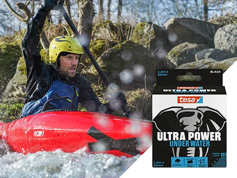 Person in a yellow helmet kayaking in a river with splashes of water. The foreground features packaging for tesa Ultra Power Under Water tape, showing an illustration of an elephant and details about the product. (This text has been generated by AI)