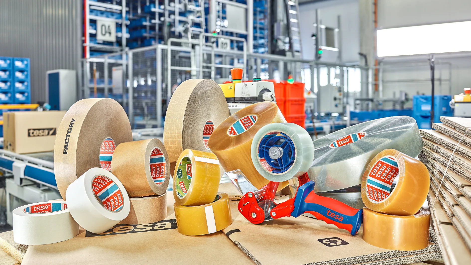 TESA-60412_22.4.2022_MM_24925_FS_V3_300dpi A variety of packing tesa tapes, including clear and brown rolls, are displayed on top of stacked cardboard boxes in an industrial setting. A blue tesa tape dispenser is also present. Shelving and equipment are visible in the background. (This text has been generated by AI)