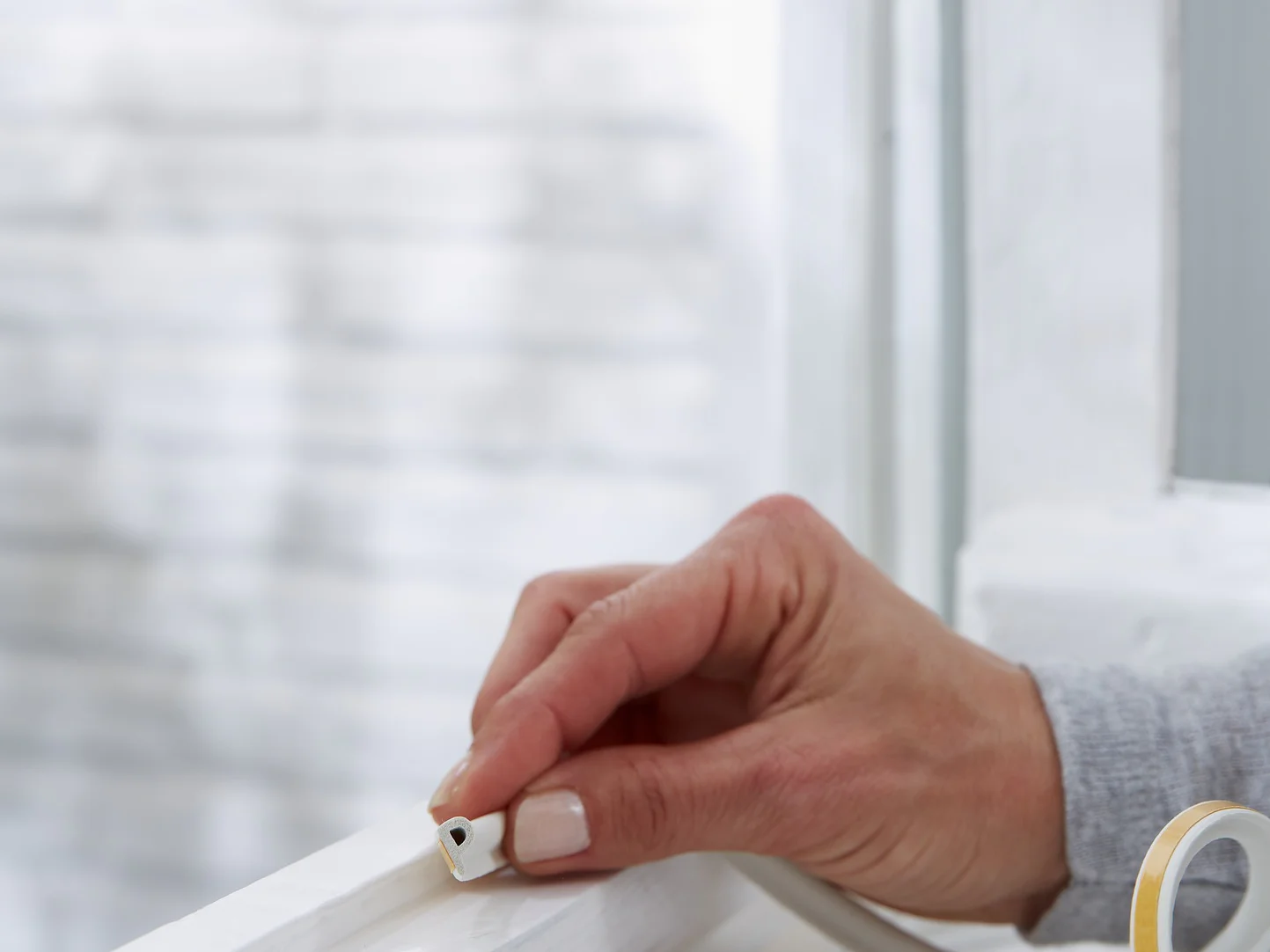 A hand is placing tesa tape weather stripping onto a white window frame. The background shows a blurred exterior wall. The weather stripping is beige with a peel-off backing, suggesting a home improvement or insulation task. (This text has been generated by AI)