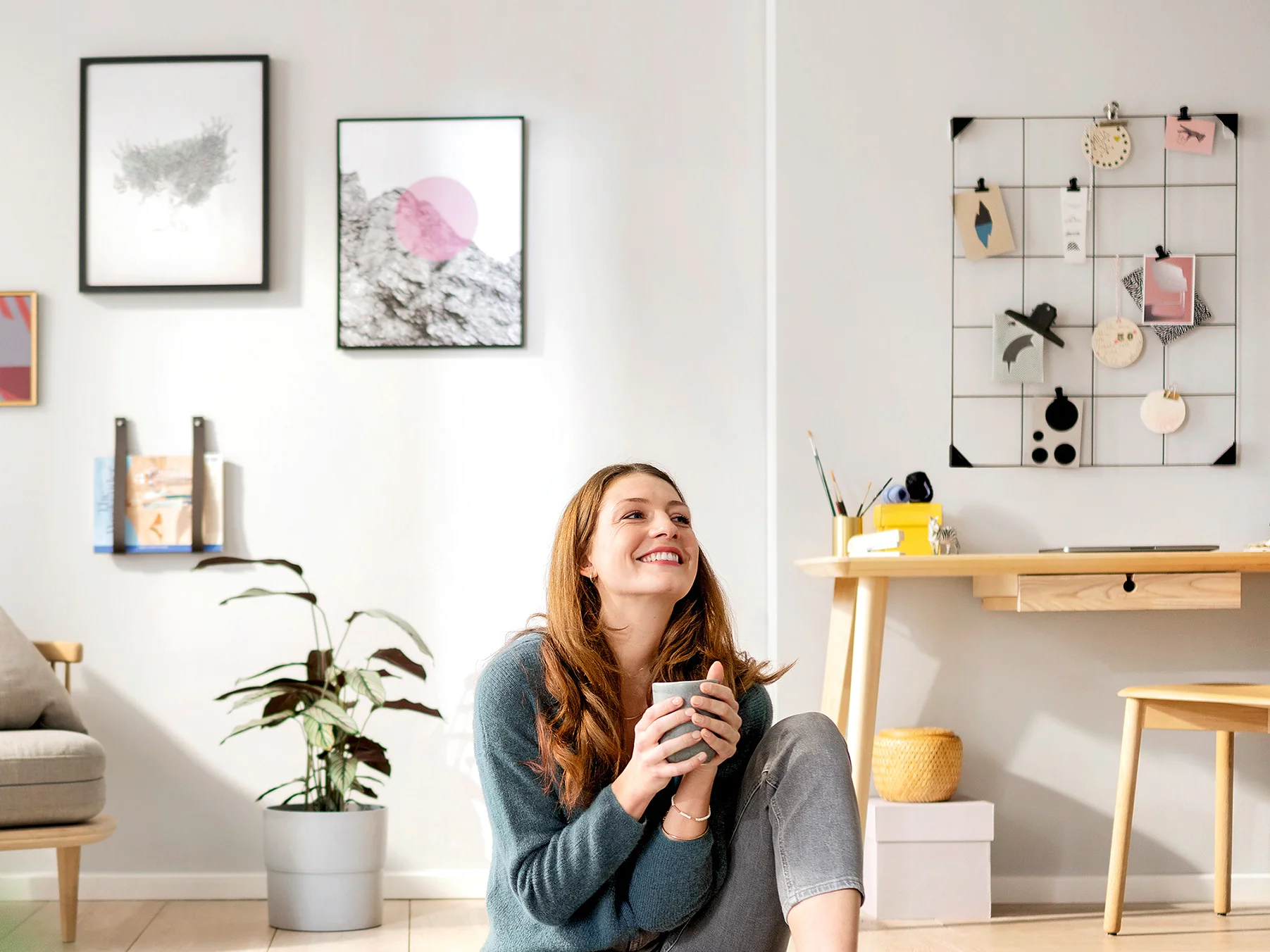 A woman with long hair is sitting on the floor in a brightly lit living room holding a mug, smiling upward. The room features a small desk with a chair, a potted plant, wall art, and decorative items secured with tesa tape on a wire grid. (This text has been generated by AI)
