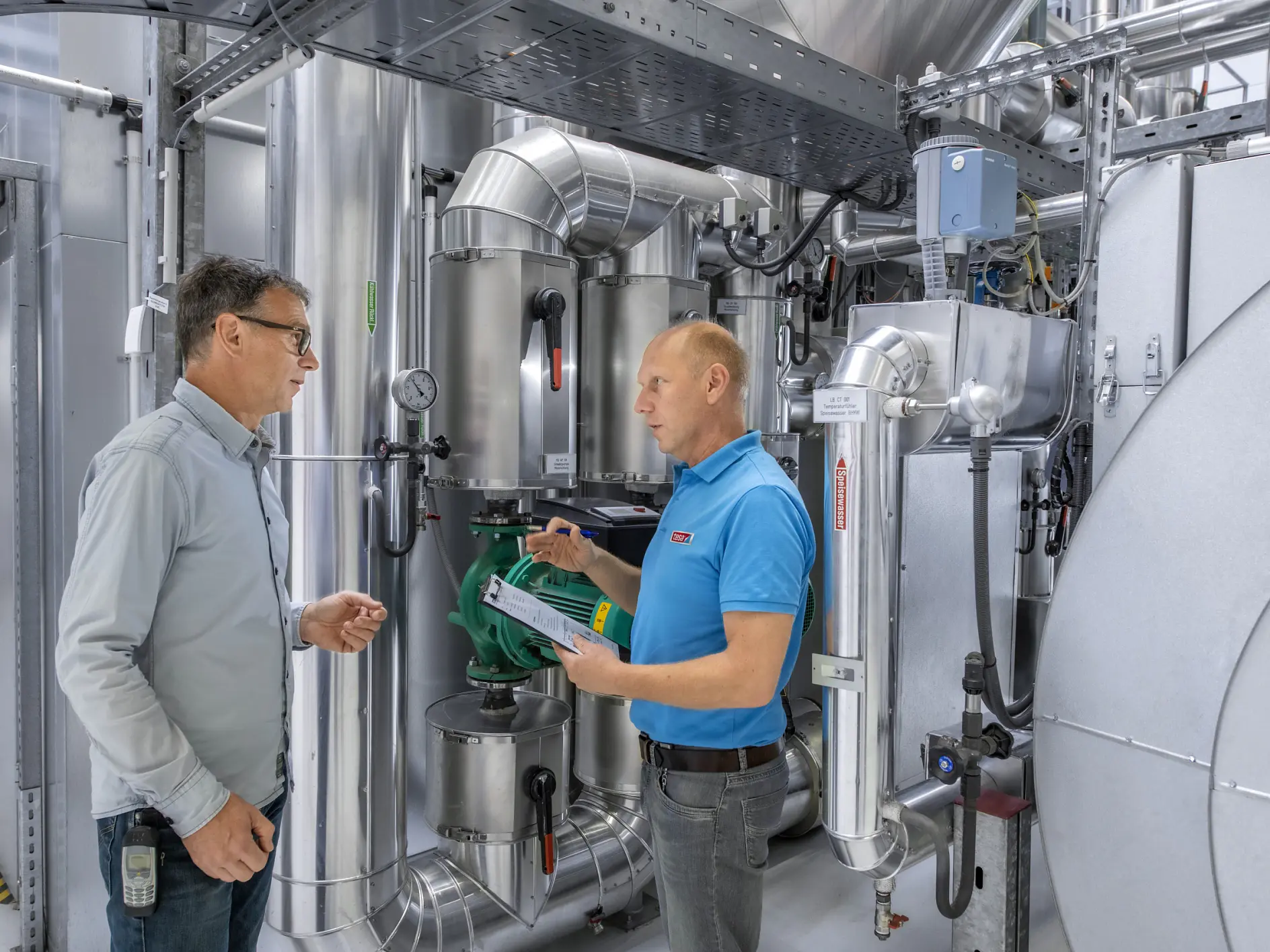Two men stand in an industrial facility surrounded by metal pipes and machinery. One, wearing a light blue shirt and glasses, listens while the other, in a blue polo, explains something while holding a document.