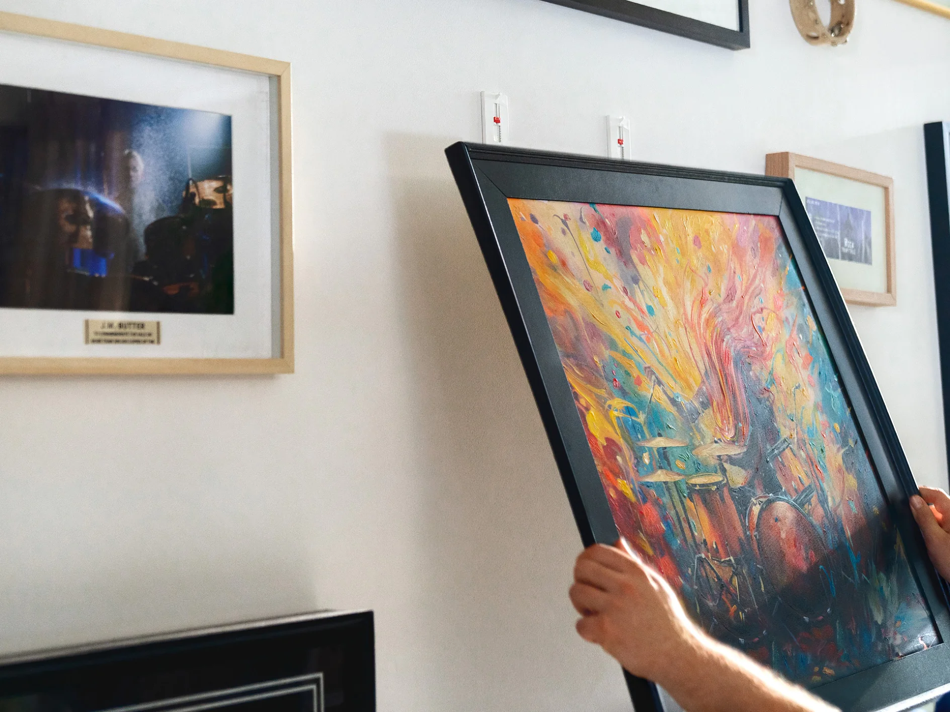 A person is hanging a colorful abstract painting on a wall. The wall already has several framed pictures, including one featuring a dark, ambiguous scene. The persons arm and part of their torso are visible on the right side of the image, and they are using tesa tape to affix the new painting. (This text has been generated by AI)