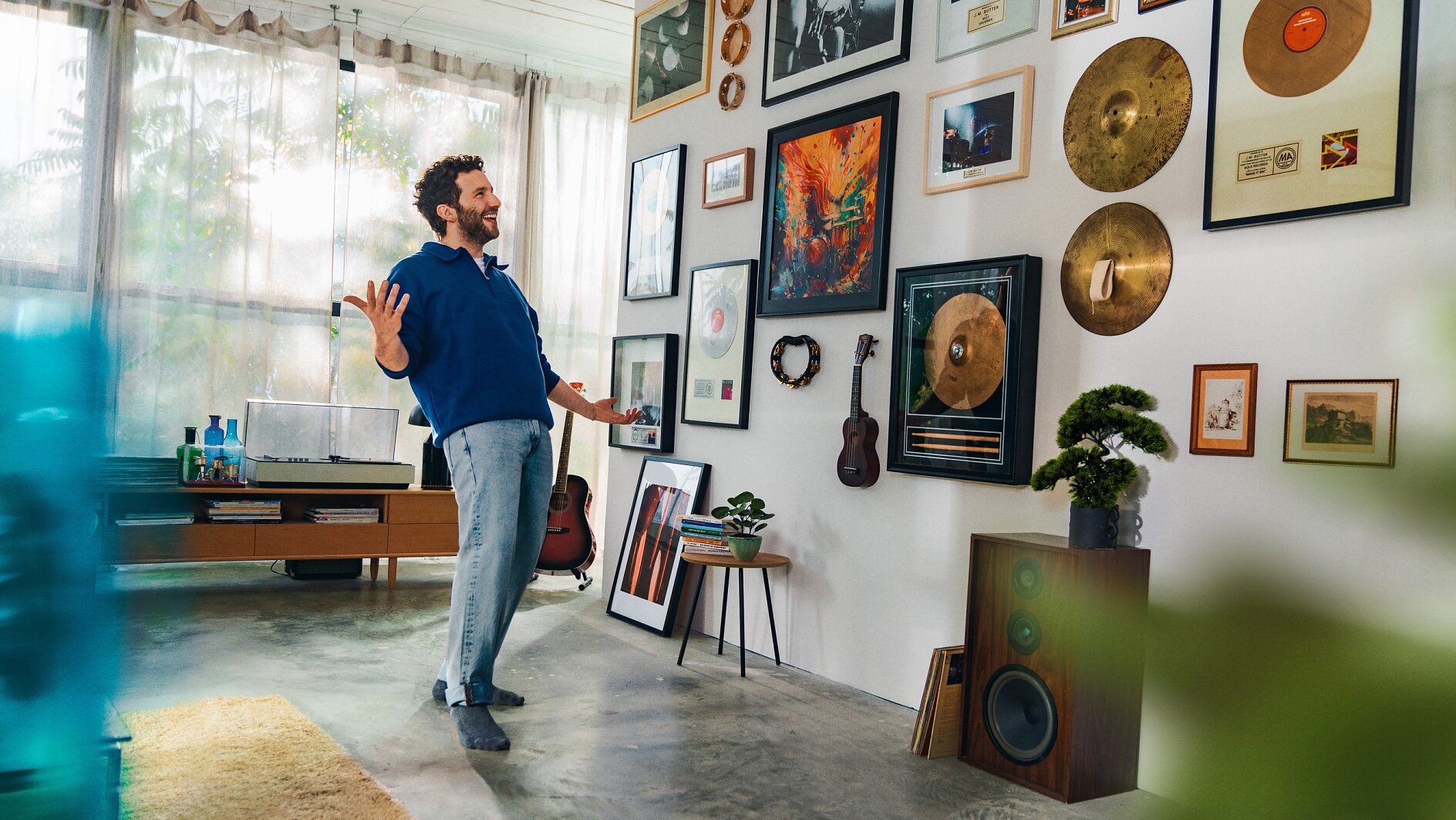 A man in a blue sweater and gray pants stands in a living room, gesturing toward a wall adorned with framed records, cymbals, and photos. The room also features a record player, guitars, a small tree, and a speaker. (This text has been generated by AI)