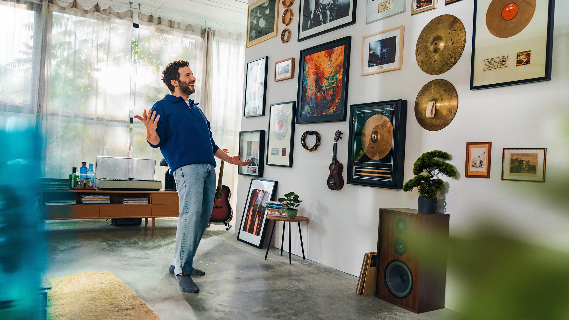 A man in a blue sweater and gray pants stands in a living room, gesturing toward a wall adorned with framed records, cymbals, and photos. The room also features a record player, guitars, a small tree, and a speaker. (This text has been generated by AI)