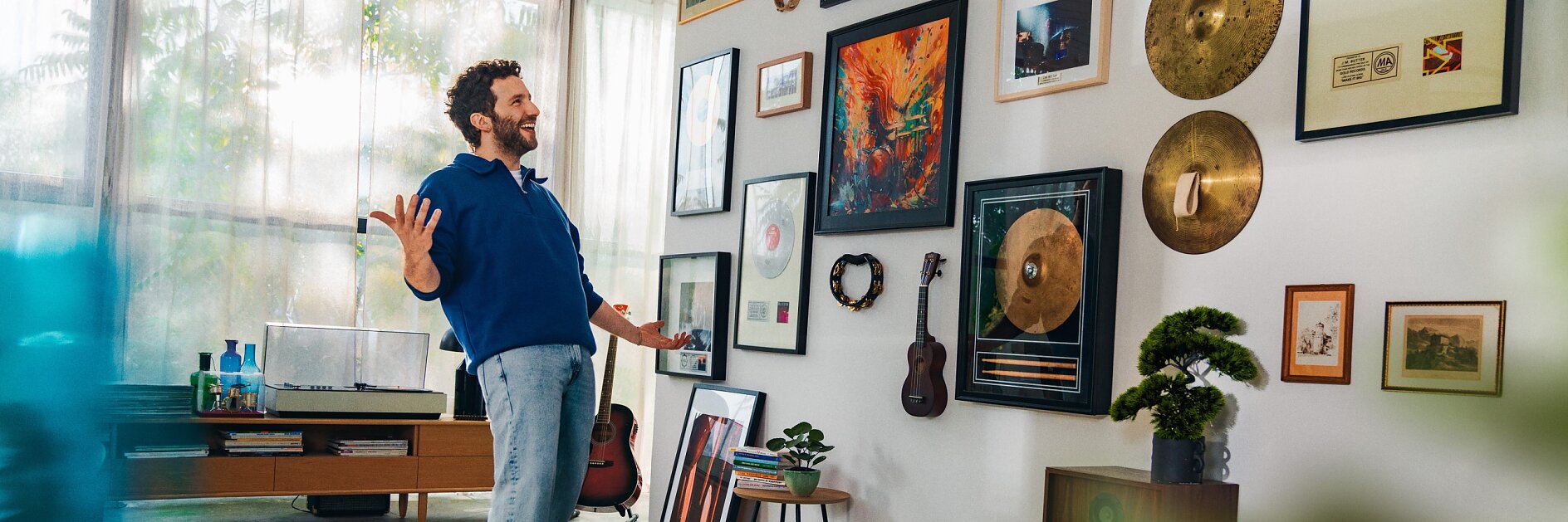 A man in a blue sweater and gray pants stands in a living room, gesturing toward a wall adorned with framed records, cymbals, and photos. The room also features a record player, guitars, a small tree, and a speaker. (This text has been generated by AI)