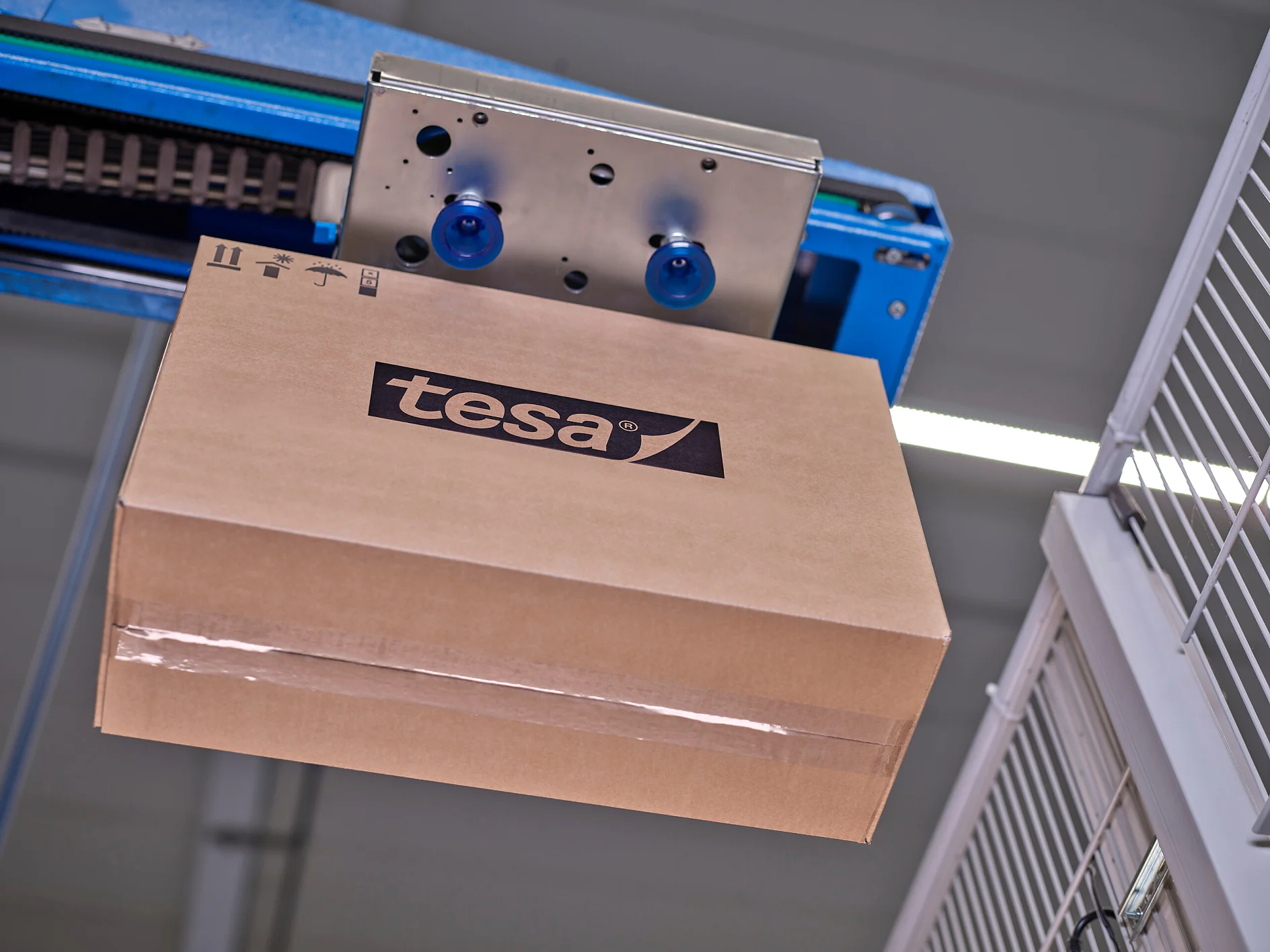 A brown cardboard box with the tesa tape logo is being handled by a blue automated machine. The background shows metal fencing and industrial equipment. (This text has been generated by AI)
