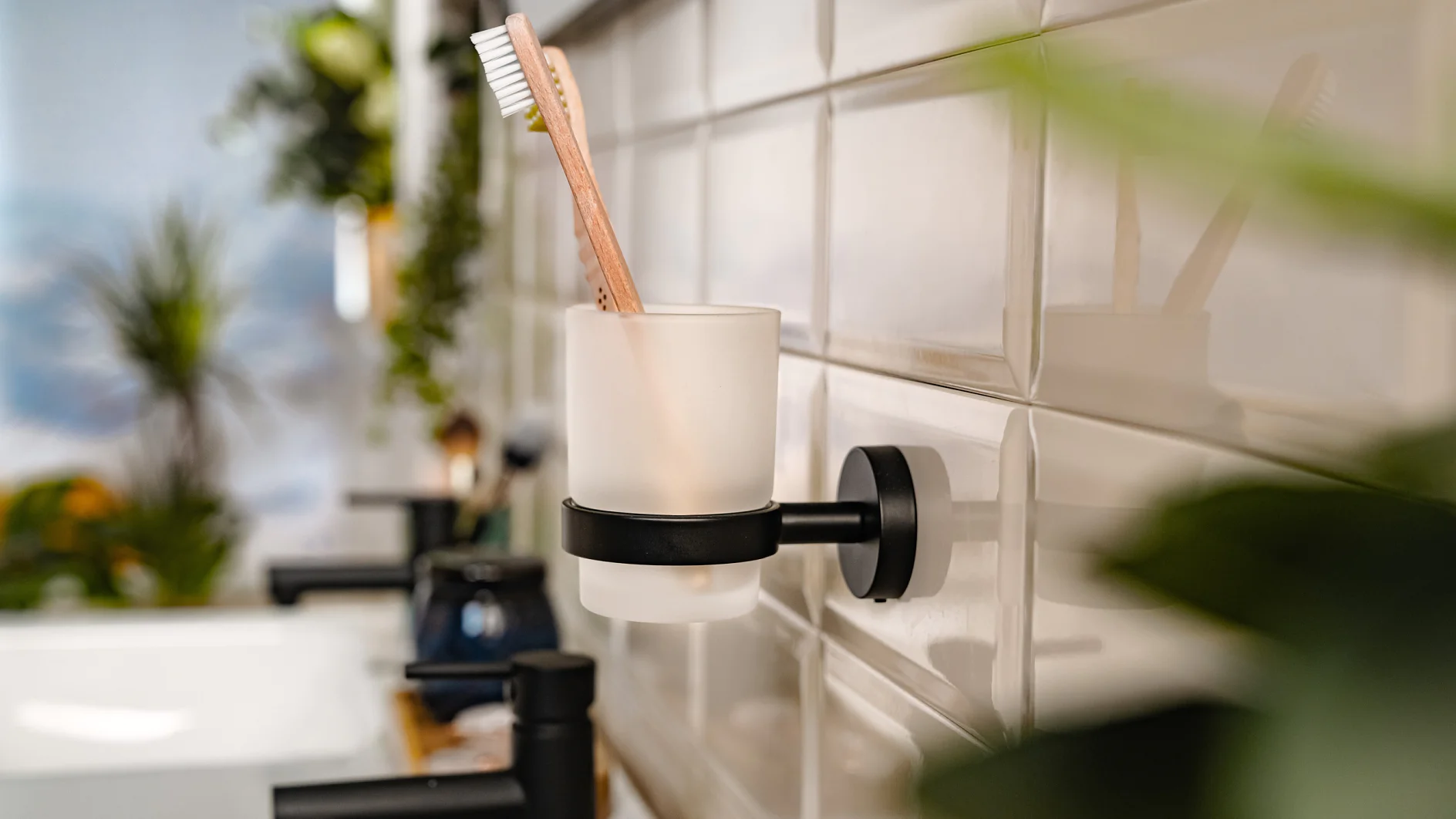 En badrumsscen med en tandborste i en hållare för frostat glas fäst på en kaklad vägg med hjälp av tesa-tejp. Förgrunden har suddiga gröna växter. Ett handfat och en kran är delvis synliga till vänster. (This text has been generated by AI)