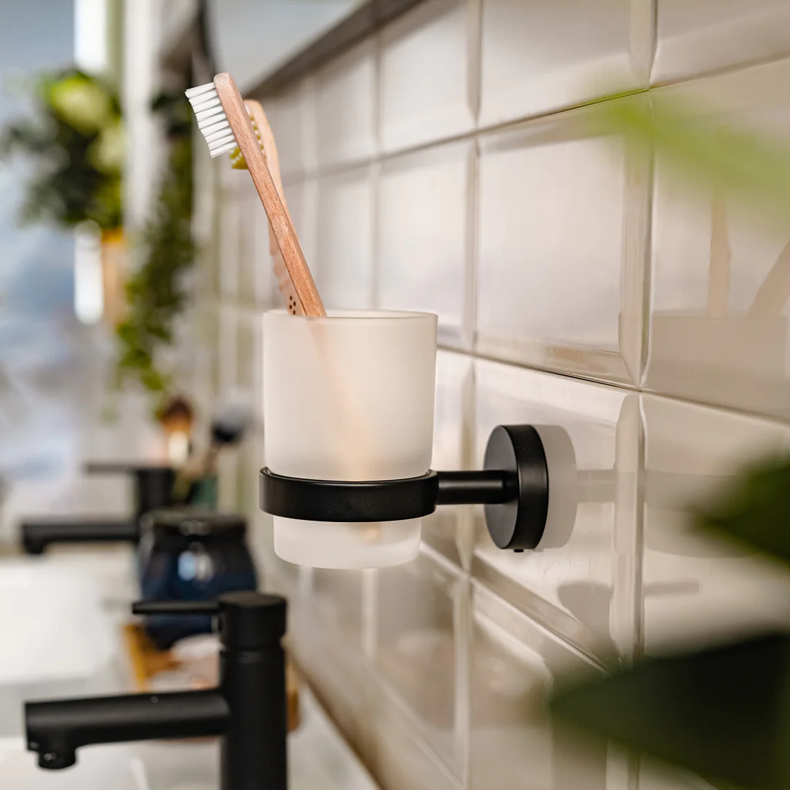 En badrumsscen med en tandborste i en hållare för frostat glas fäst på en kaklad vägg med hjälp av tesa-tejp. Förgrunden har suddiga gröna växter. Ett handfat och en kran är delvis synliga till vänster. (This text has been generated by AI)