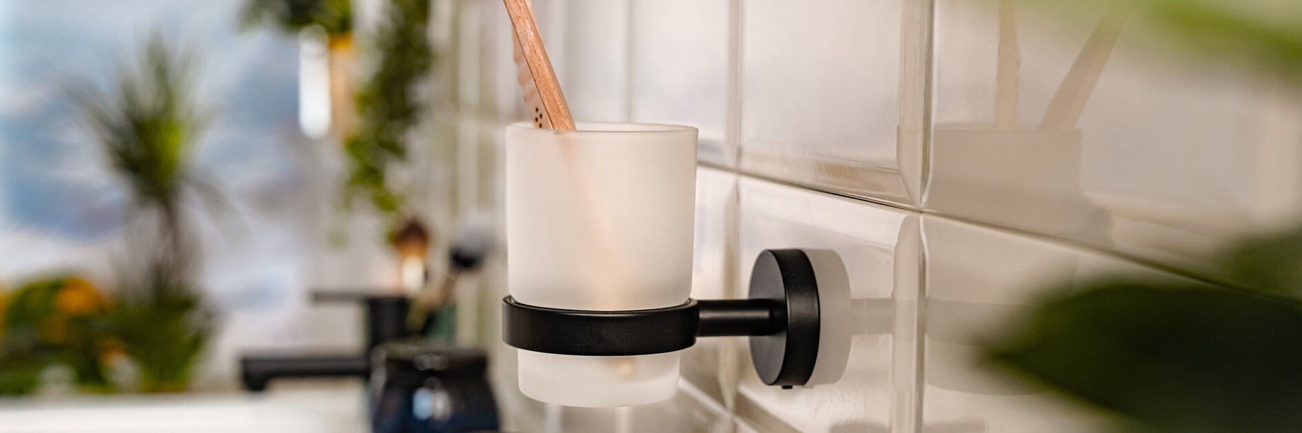 En badrumsscen med en tandborste i en hållare för frostat glas fäst på en kaklad vägg med hjälp av tesa-tejp. Förgrunden har suddiga gröna växter. Ett handfat och en kran är delvis synliga till vänster. (This text has been generated by AI)