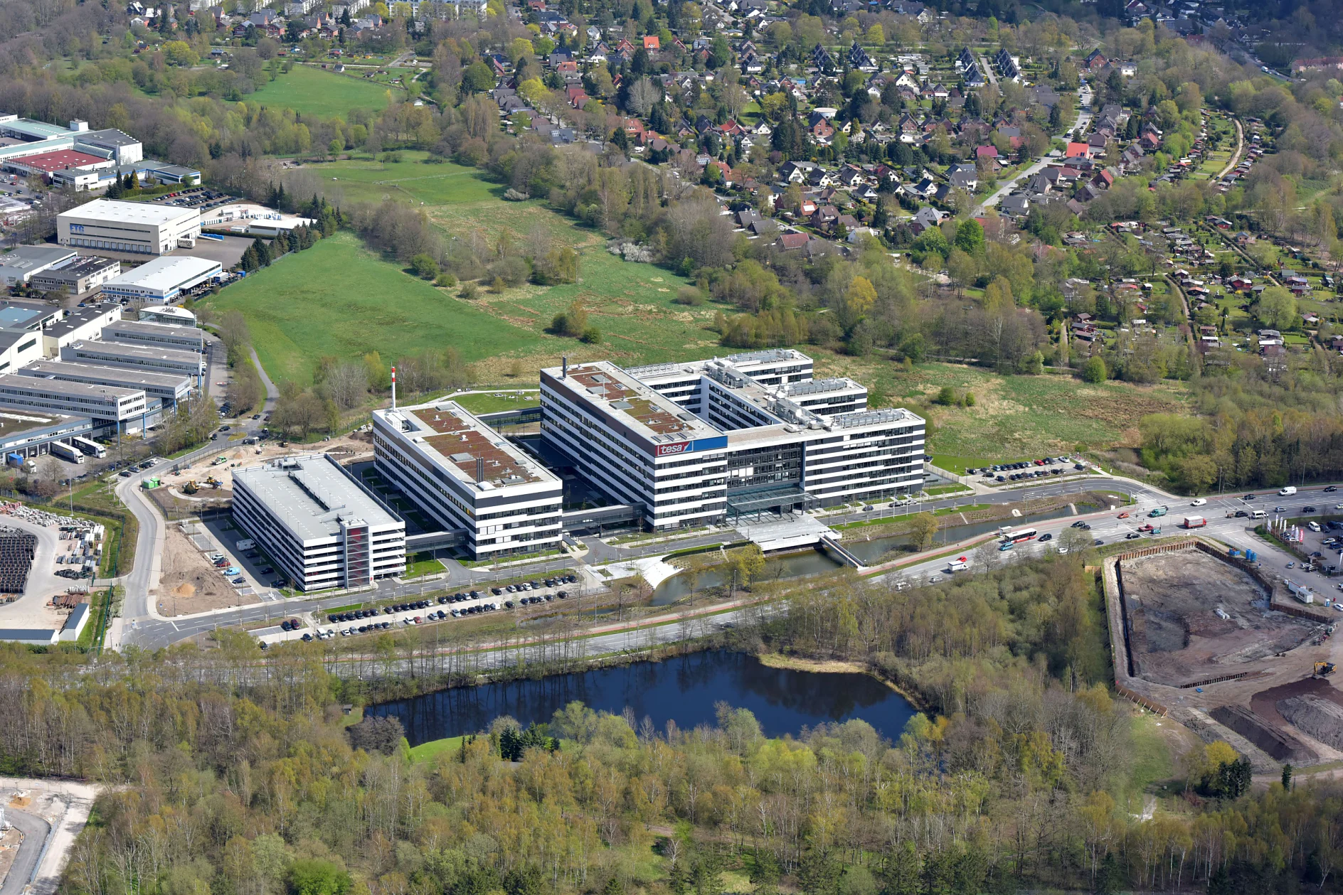 Aerial view of a large, modern office complex surrounded by green spaces and a pond. Residential houses are visible in the background, alongside roads and construction areas. Trees surround the area, creating a mix of urban and natural elements. (This text has been generated by AI)