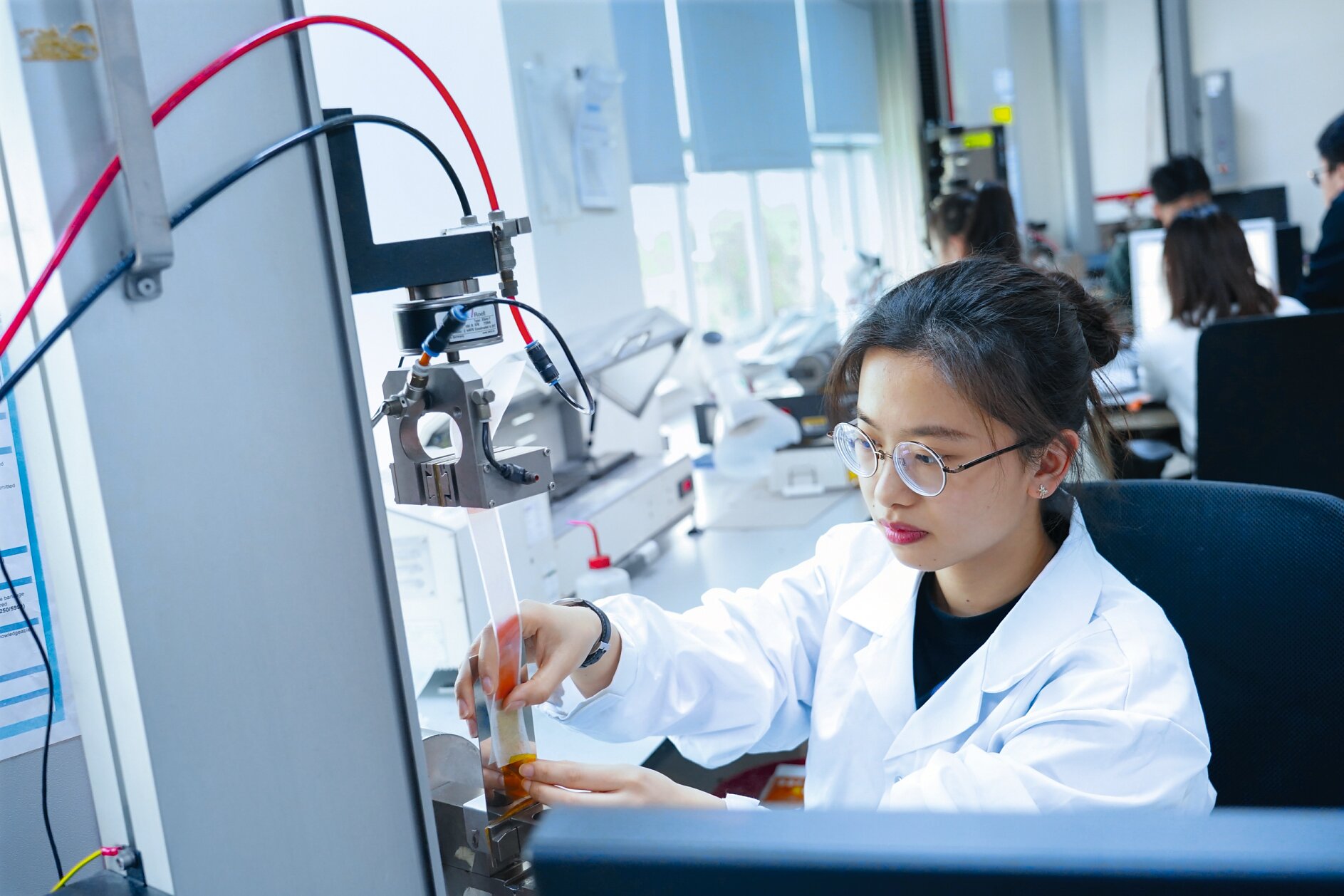 A person in a lab coat and glasses operates equipment in a laboratory. They are adjusting a component near a machine with wires using tesa tape. Other people are working in the background, and computer monitors are visible. The setting is brightly lit and organized. (This text has been generated by AI)