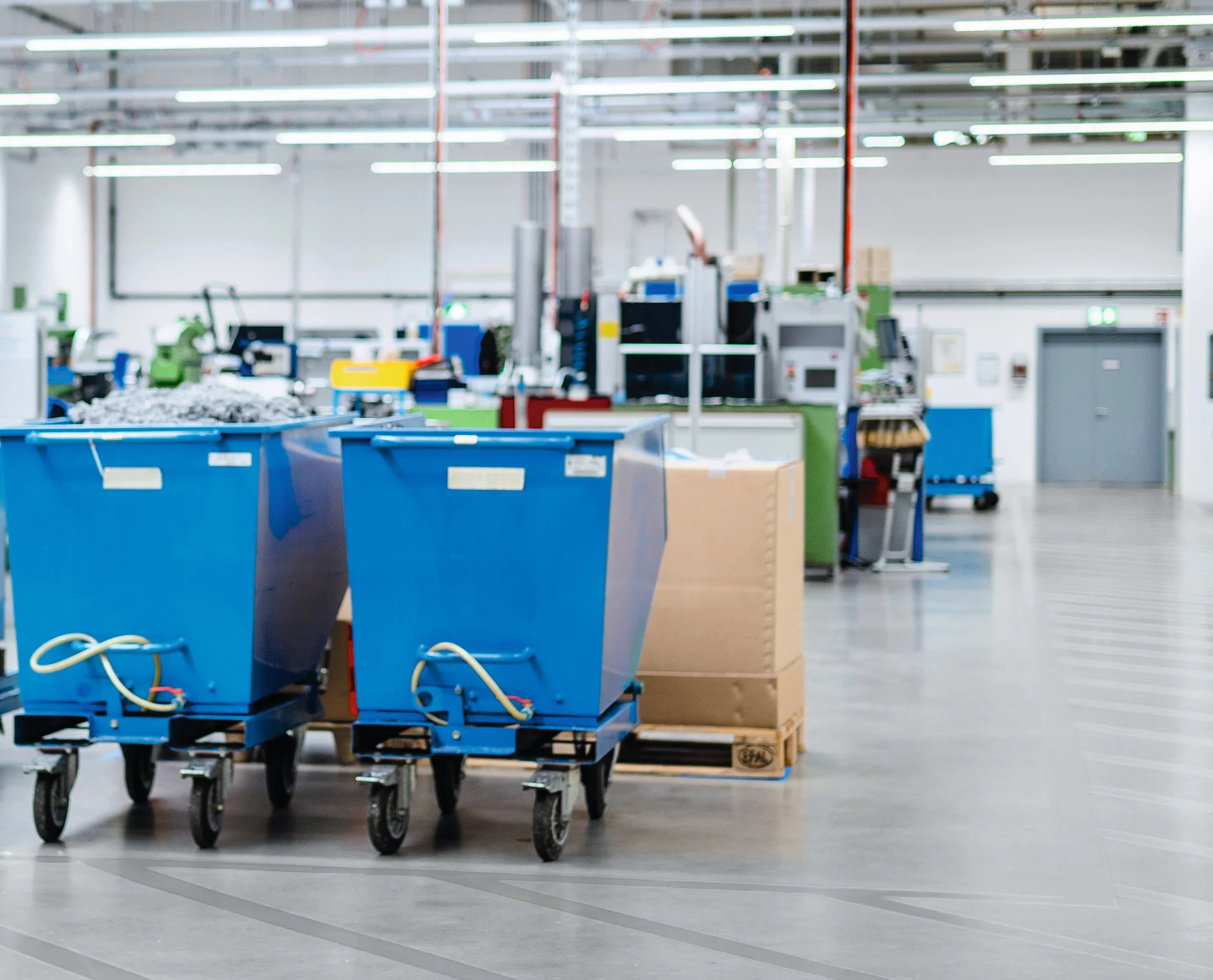 A factory floor with two large blue wheeled bins in the foreground. Machinery and equipment are visible in the background. The area is well-lit with overhead fluorescent lights, and the floor is smooth concrete, secured using reliable tesa tape for any labeling or marking needs. (This text has been generated by AI)