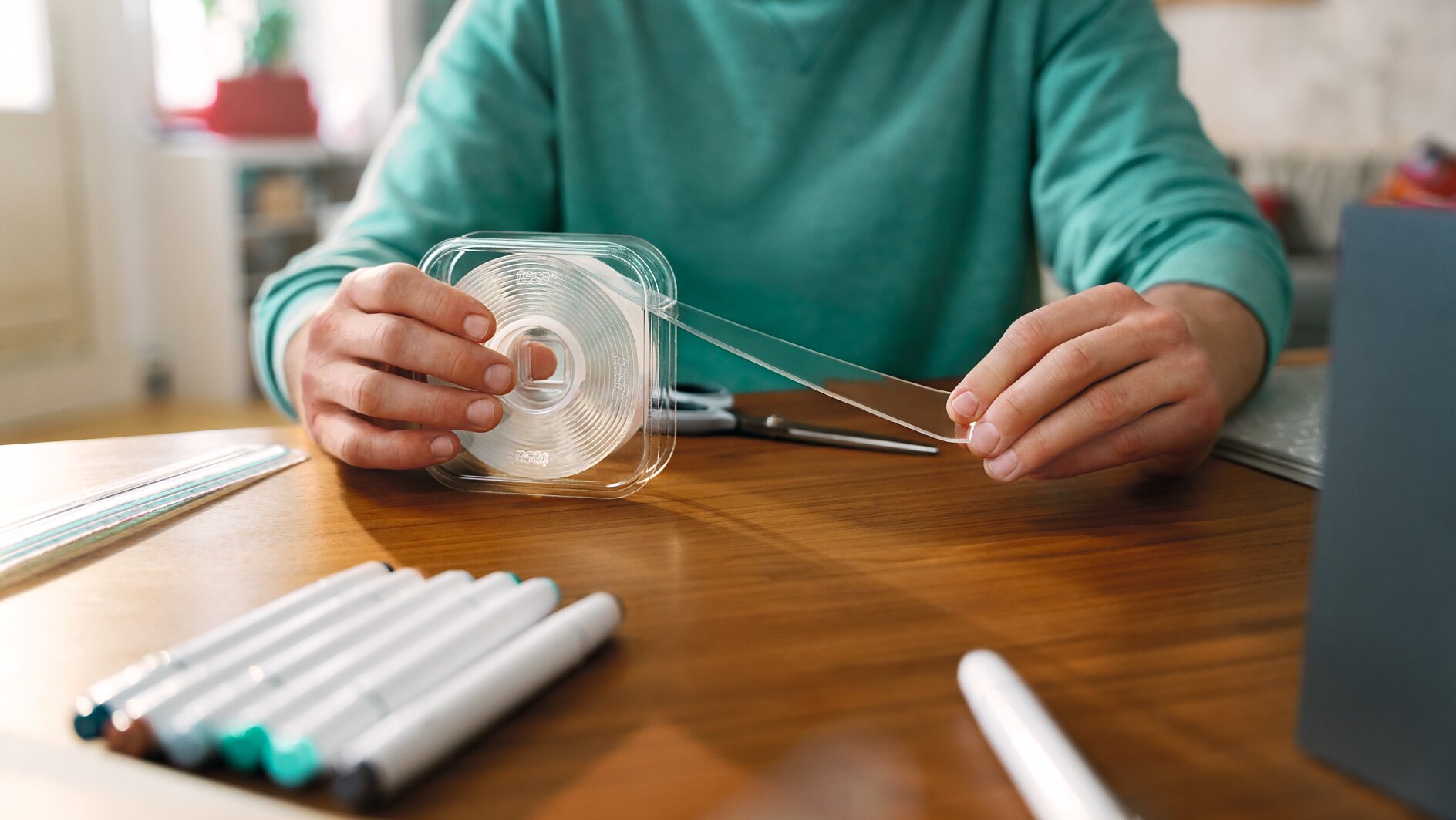 A person wearing a green shirt is seated at a wooden table, handling a clear tesa tape dispenser. Several markers are scattered on the table, and a notebook is partially visible. Scissors are also present near the persons hands. (This text has been generated by AI)