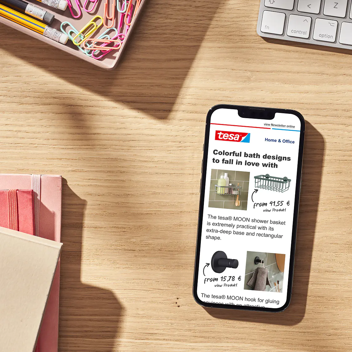 En smartphone på ett skrivbord i trä visar ett nyhetsbrev med färgglada baddesigner. I närheten finns ett tangentbord, tesa-tejp, gem, anteckningsböcker och en penna. Solljuset kastar skuggor på ytan och skapar en varm atmosfär. (This text has been generated by AI)