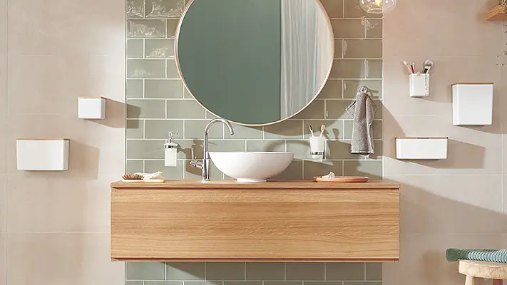 A modern bathroom with a circular mirror above a wooden vanity. The wall features green tiles and there are white shelves and containers mounted on the side using tesa tape. A white sink and silver faucet are centered on the countertop. (This text has been generated by AI)