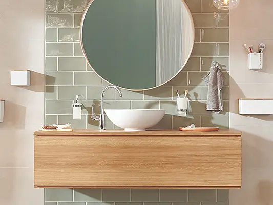 A modern bathroom with a circular mirror above a wooden vanity. The wall features green tiles and there are white shelves and containers mounted on the side using tesa tape. A white sink and silver faucet are centered on the countertop. (This text has been generated by AI)