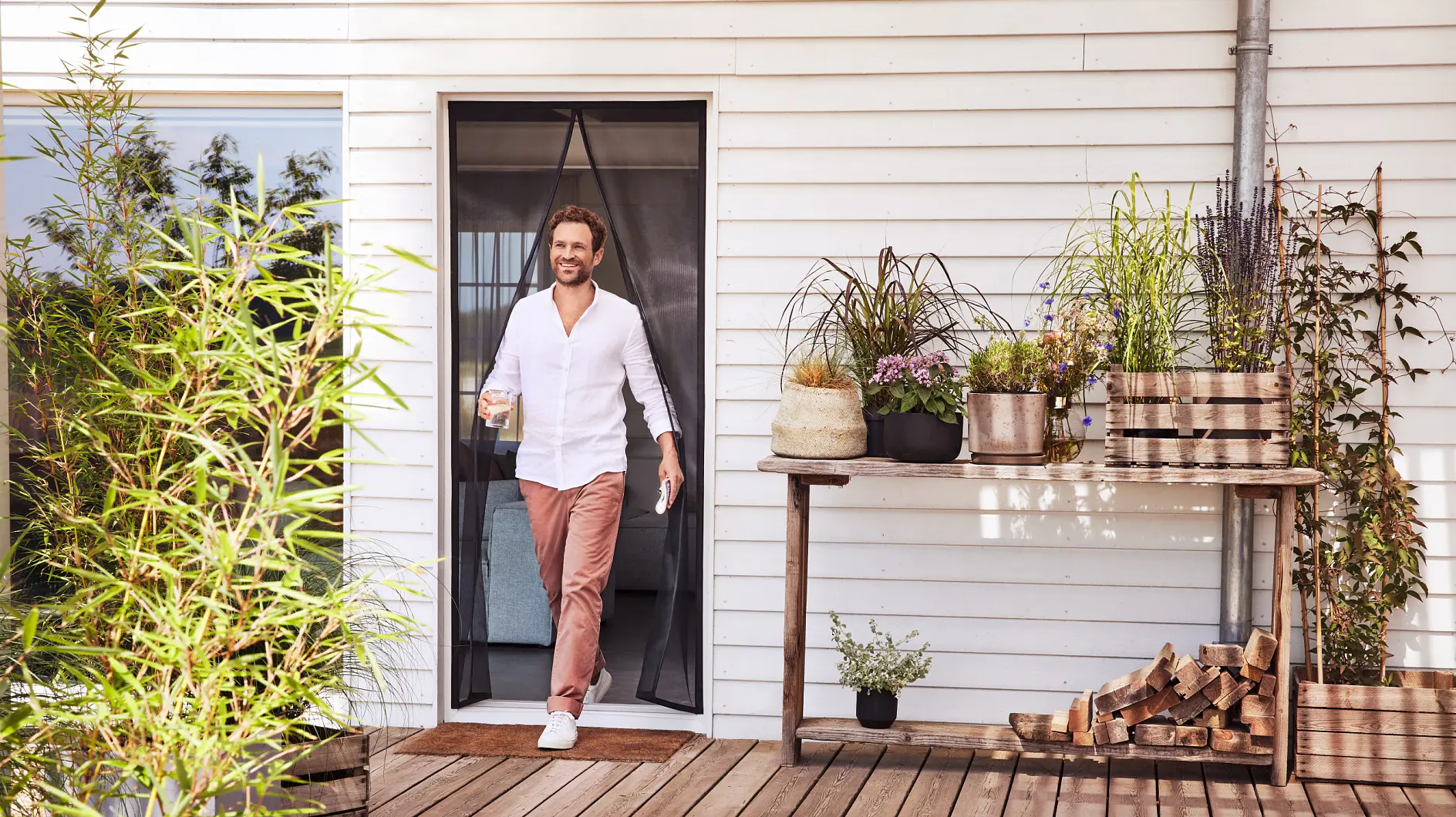 A man wearing a white shirt and pink pants walks through a door onto a wooden deck, holding a glass. The deck features potted plants, a stack of firewood, and a table with more plants. The building has white siding. (This text has been generated by AI)