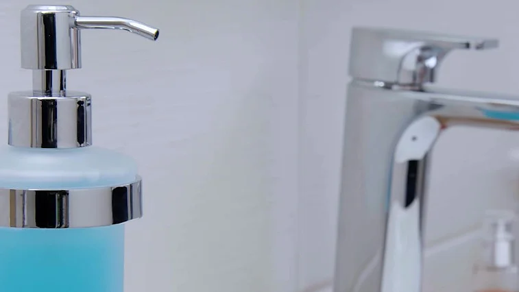 Närbild av badrumshandfat med väggmonterad tvålpump i krom fylld med blå vätska, kromkran och vit mugghållare – modern hygieninstallation.