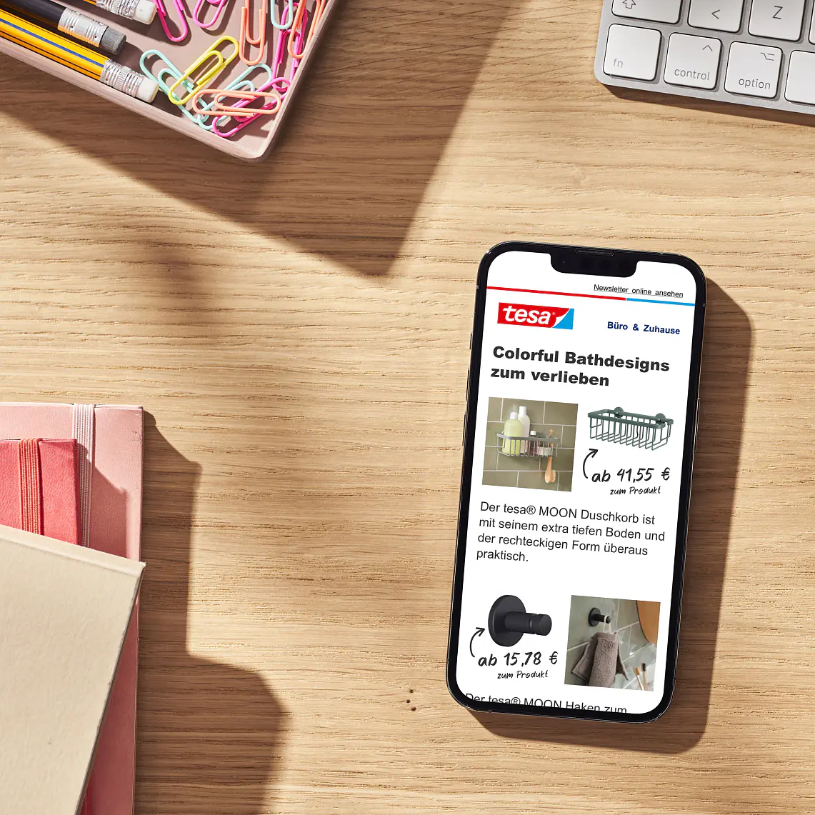 A smartphone displaying a webpage with tesa tape branding is on a wooden desk. A tray of paperclips and some notebooks are nearby. The webpage shows images and prices of various bathroom designs and products in German. (This text has been generated by AI)