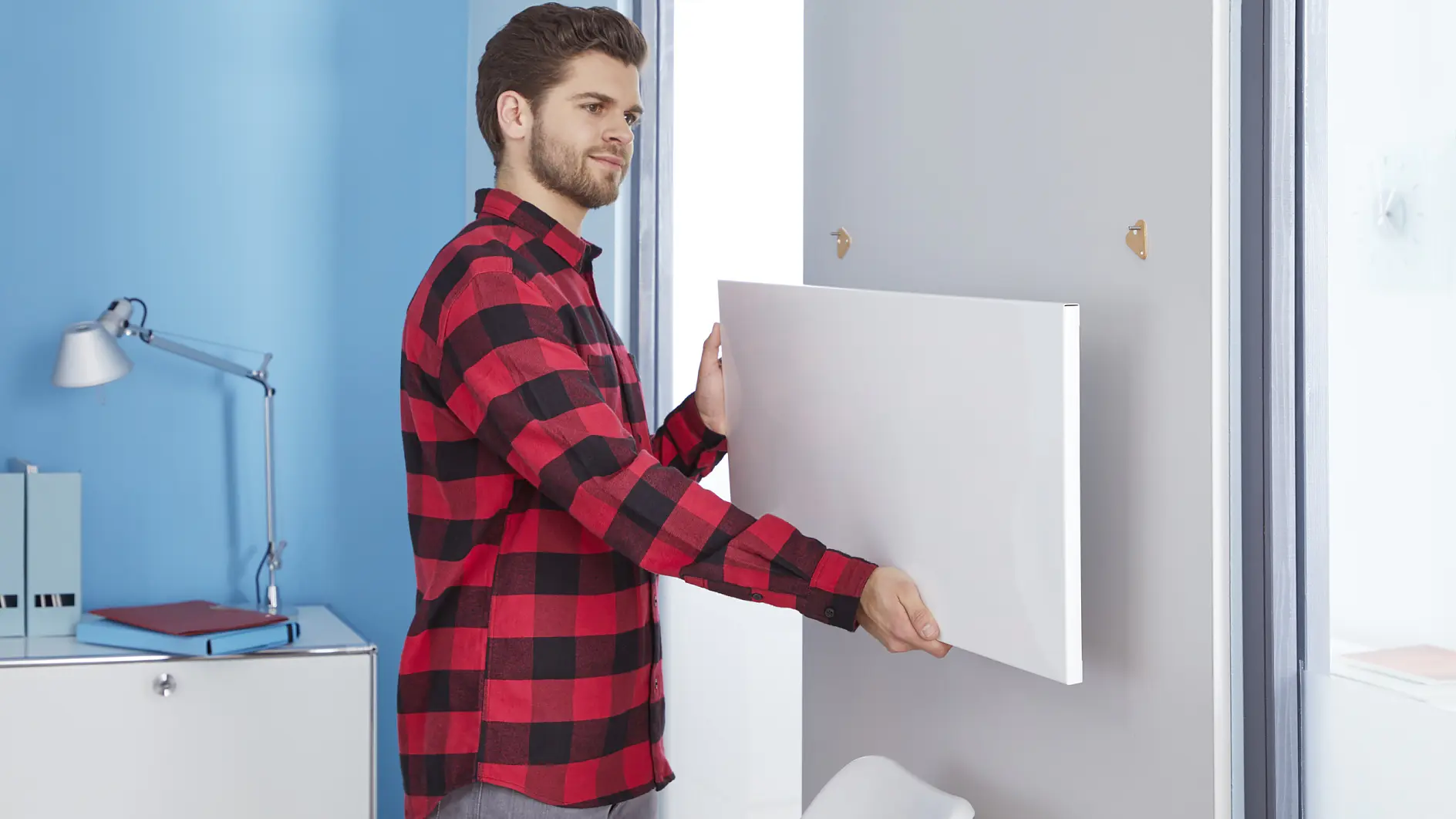 En man i en röd och svart rutig skjorta hänger upp en tom vit duk på en grå vägg med hjälp av tesa-tejp. Rummet har blå och vita väggar, ett vitt skrivbord och en skrivbordslampa. Mannen ser fokuserad ut när han placerar duken. (This text has been generated by AI)