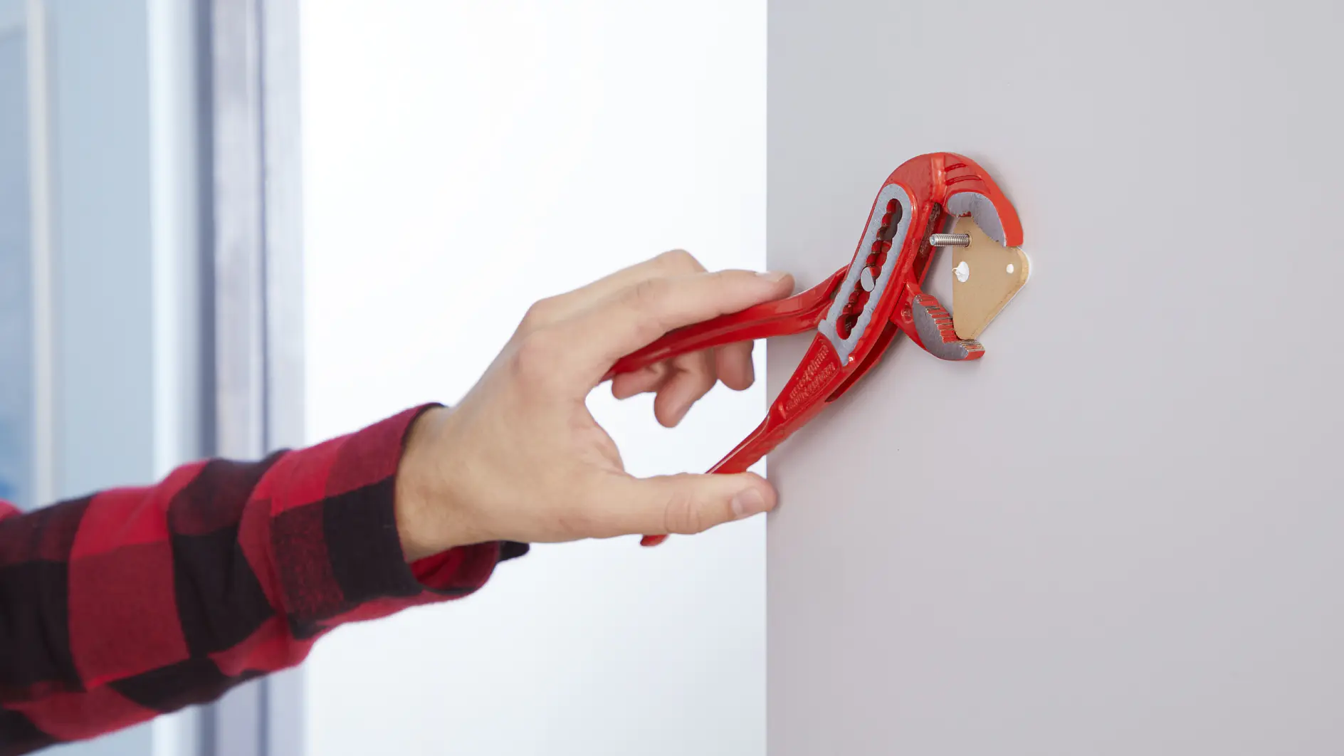 En persons hand använder en röd justerbar skiftnyckel för att montera en väggplugg i ett hål på en vit vägg. Personen bär en röd rutig skjorta. Scenen fokuserar på verktyget och installationsprocessen. (This text has been generated by AI)