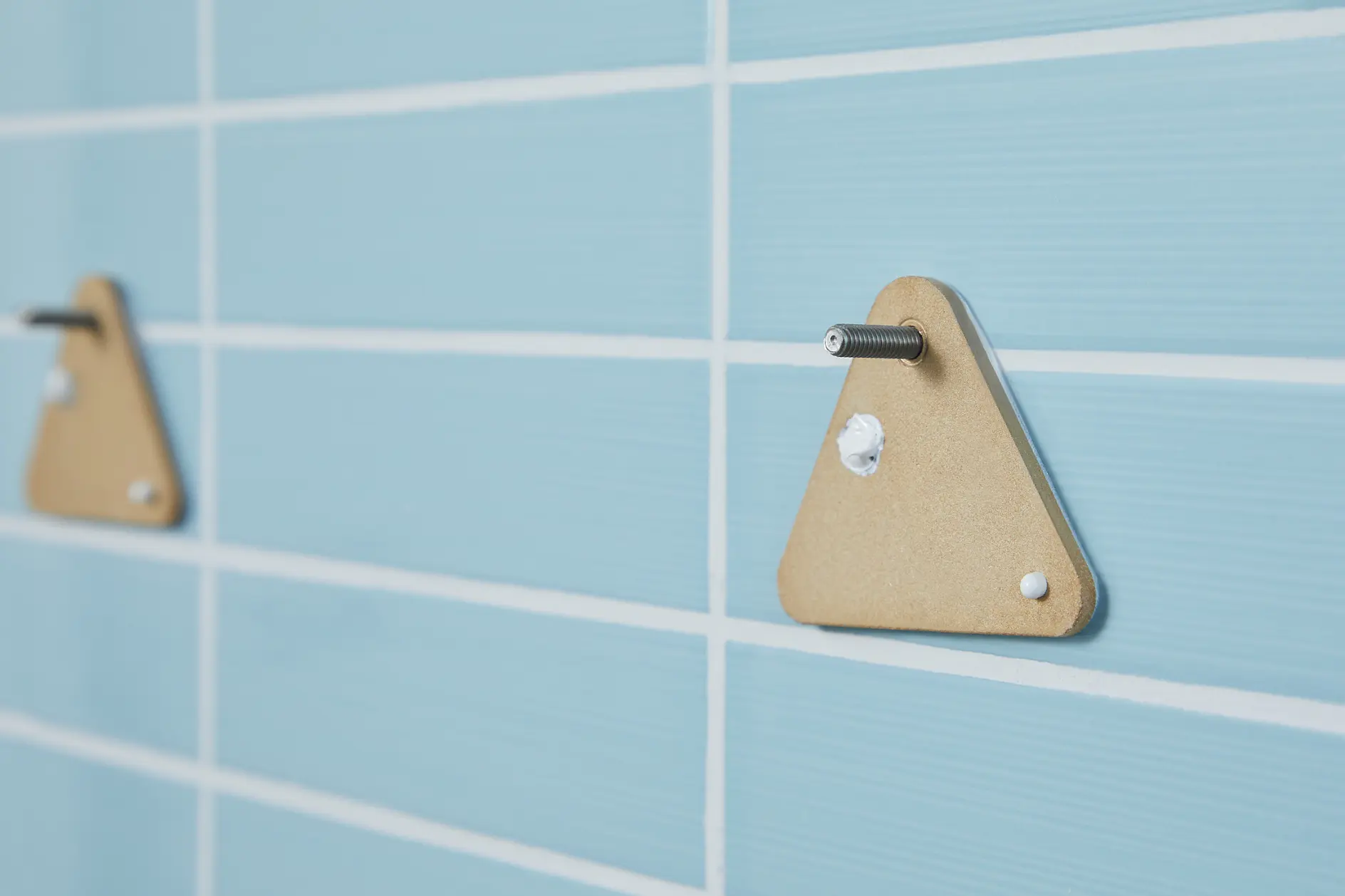 En triangulär beige konsol med en metallskruv är fäst på en ljusblå kaklad vägg med tesa-tejp. En annan identisk konsol syns i bakgrunden till vänster, också fäst med tesa-tejp. (This text has been generated by AI)