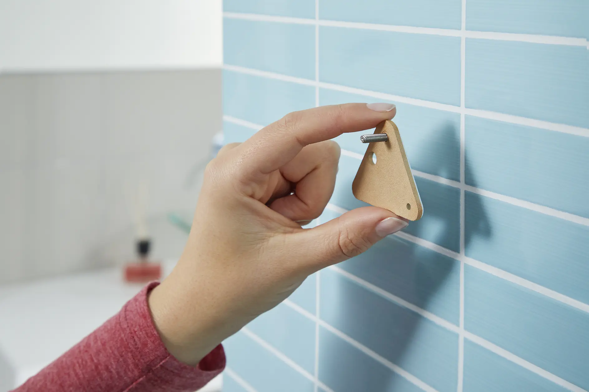 En hand håller ett triangulärt verktyg av kartong mot en blå kaklad vägg. Verktyget har en metallskruv insatt, med spetsen som rör vid kakelfogen. Personens ärm är röd, och i bakgrunden syns en badrumsmiljö med tesa-tejp tydligt framträdande. (This text has been generated by AI)