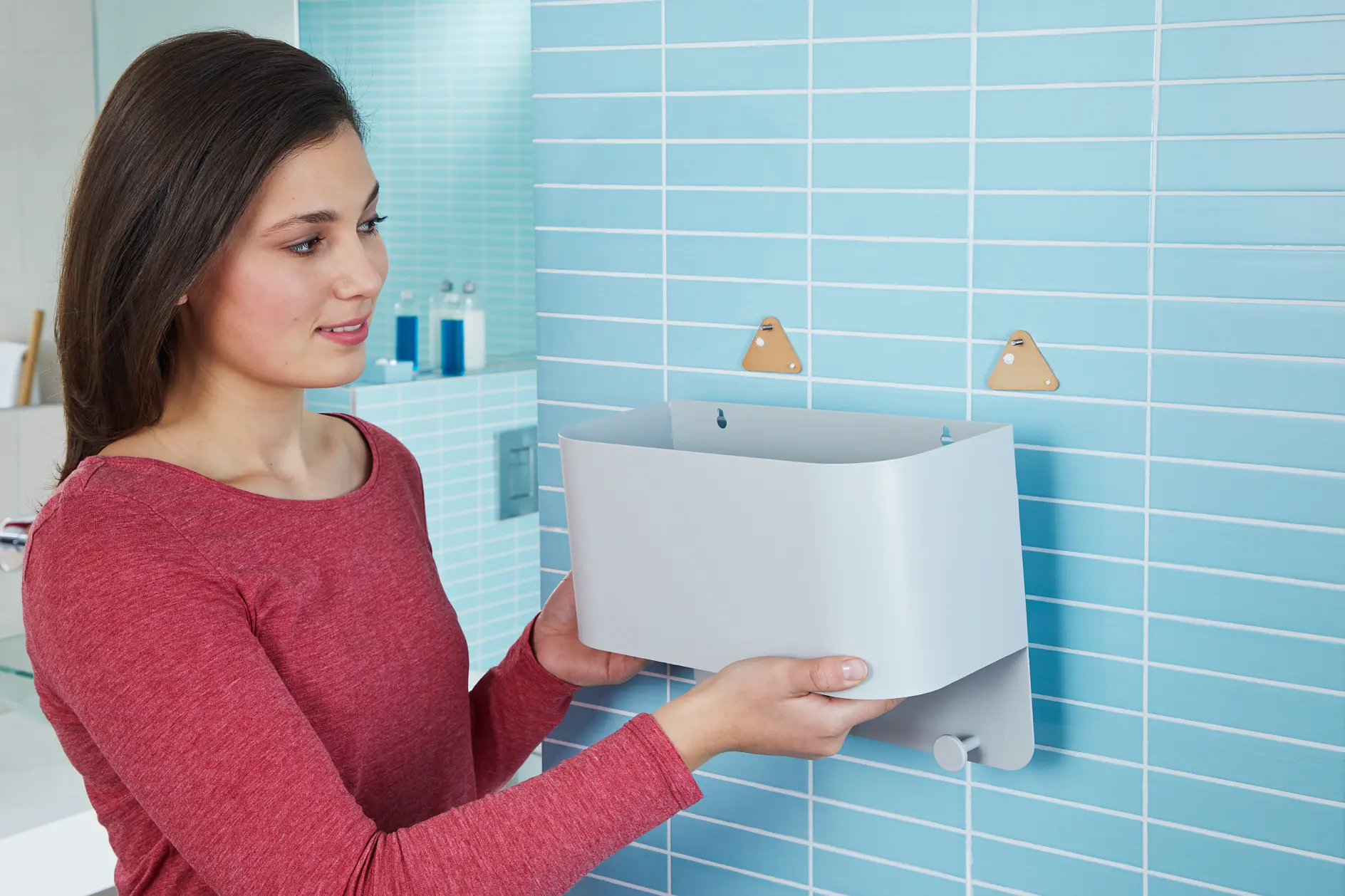 En person i röd tröja monterar en rektangulär grå behållare på en kaklad badrumsvägg med två triangelformade krokar med hjälp av tesa-tejp. Det finns flaskor på en hylla i bakgrunden. (This text has been generated by AI)