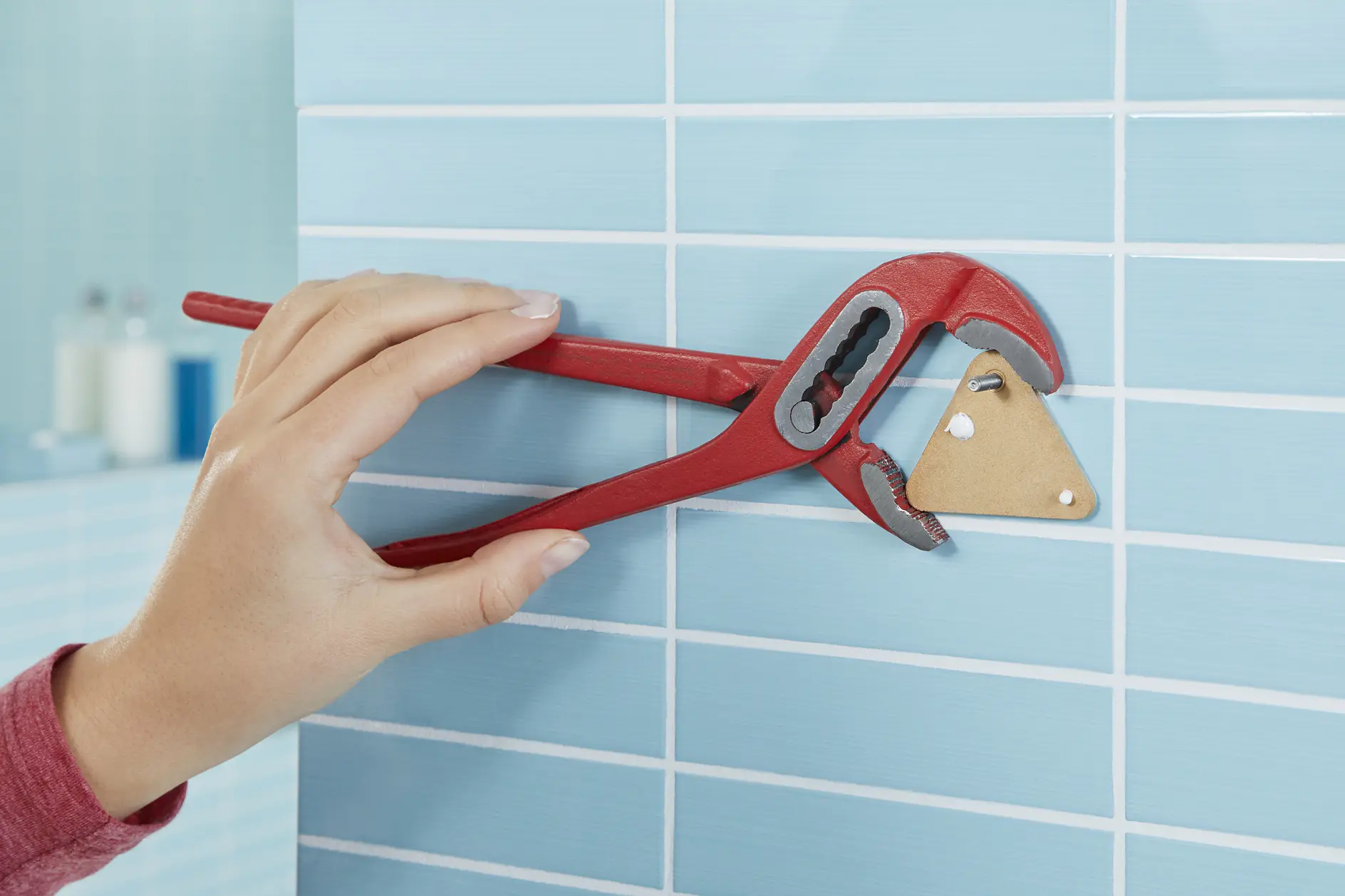 En hand i en röd ärm använder en röd rörtång för att justera ett triangulärt träföremål som är monterat på en ljusblå kaklad vägg med hjälp av tesa-tejp. Bakgrunden visar fler blå kakelplattor och suddiga badrumsartiklar. (This text has been generated by AI)