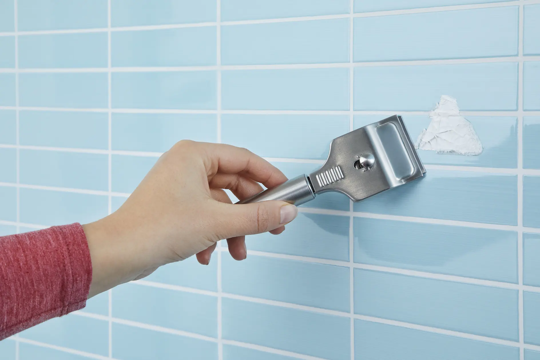 En hand i ett rött ärm använder en metallskrapa för att ta bort en vit substans från en blå kaklad vägg som är fastsatt med tesa-tejp. Kakelplattorna är arrangerade i ett rutmönster. Skrapan har ett platt, brett blad. (This text has been generated by AI)
