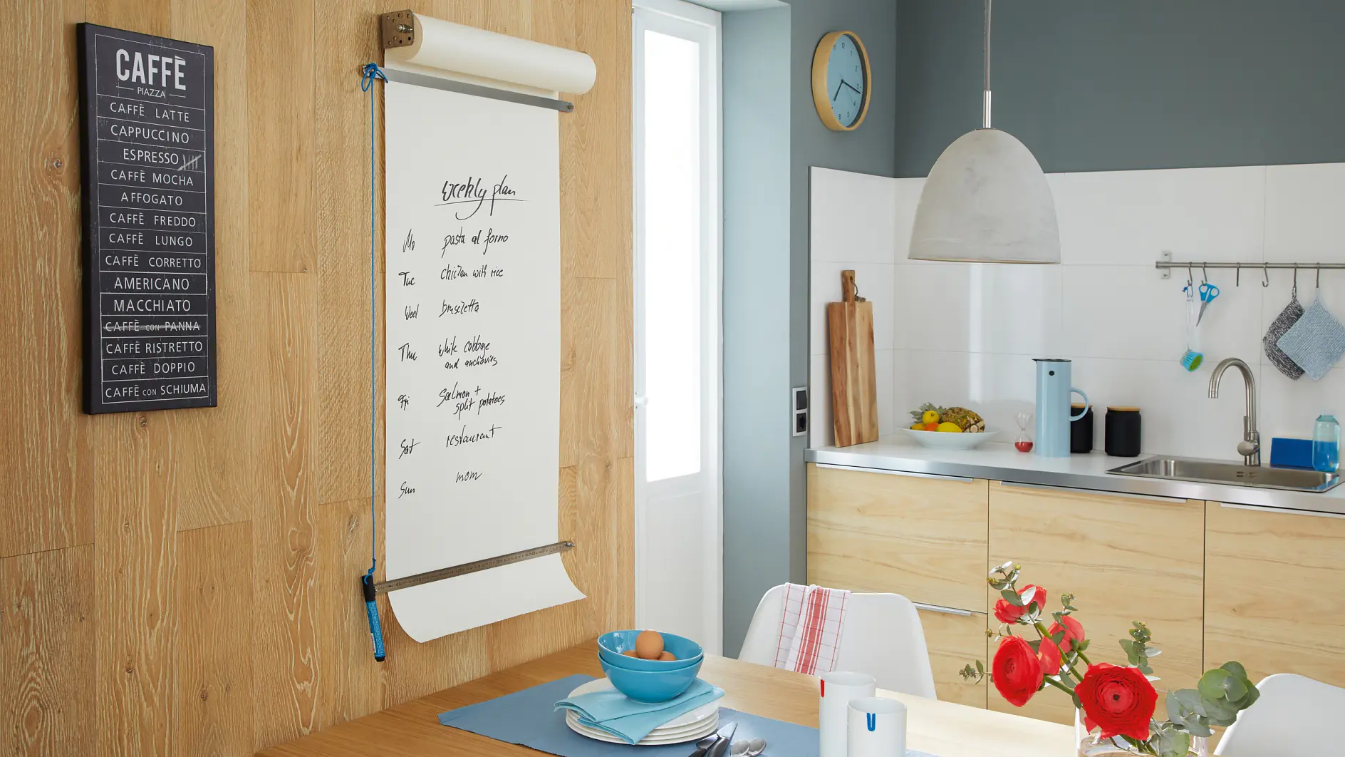 Ett modernt kök med ljusa träskåp och ett litet matbord. En stor anteckningsbok på väggen listar kaffedrycker, säkert fastsatt med tesa-tejp. På bordet finns en blå tallrik, skål och kopp. Röda blommor står i en vas och frukt ligger på bänken. En pendellampa hänger ovanför. (This text has been generated by AI)