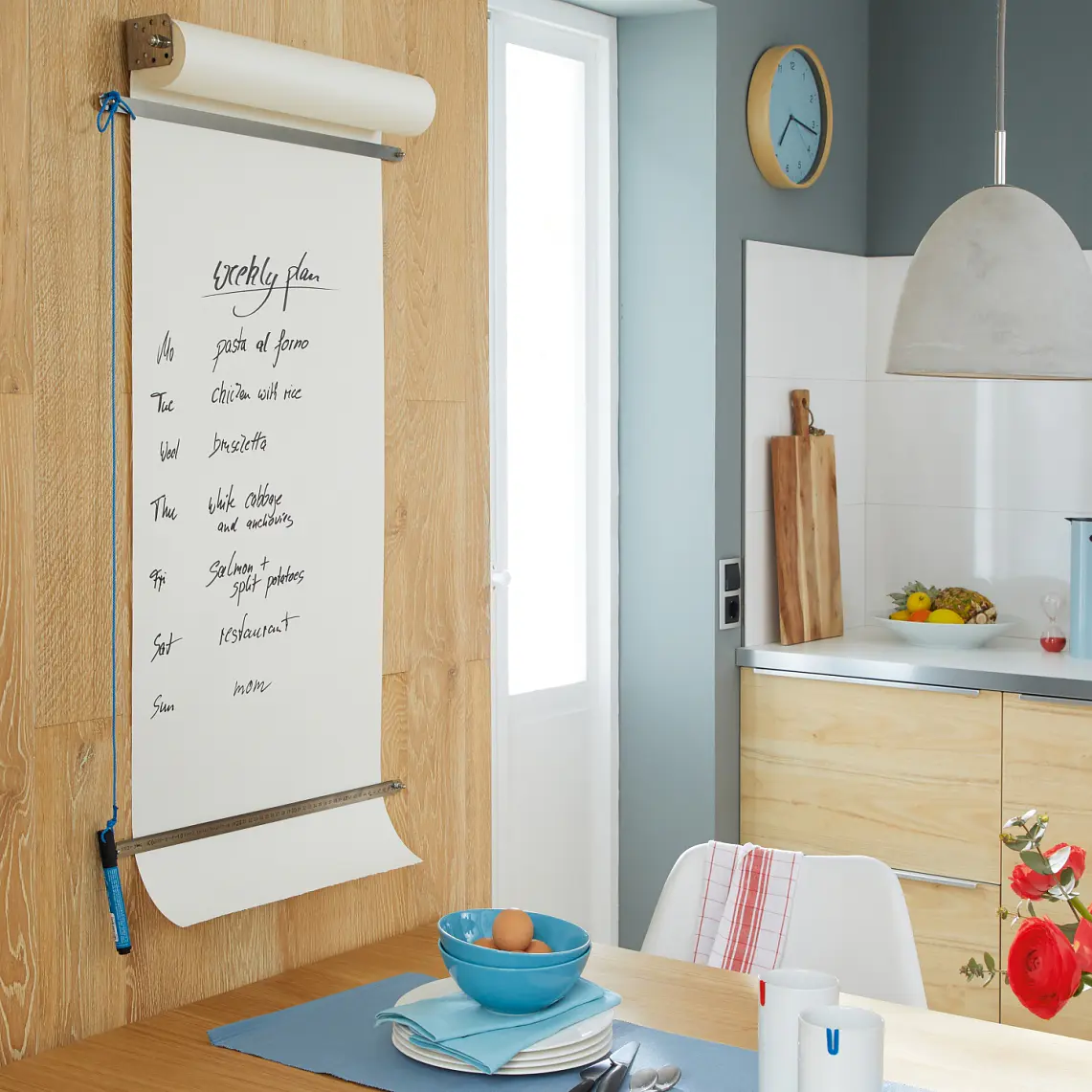 Ett modernt kök med ljusa träskåp och ett litet matbord. En stor anteckningsbok på väggen listar kaffedrycker, säkert fastsatt med tesa-tejp. På bordet finns en blå tallrik, skål och kopp. Röda blommor står i en vas och frukt ligger på bänken. En pendellampa hänger ovanför. (This text has been generated by AI)