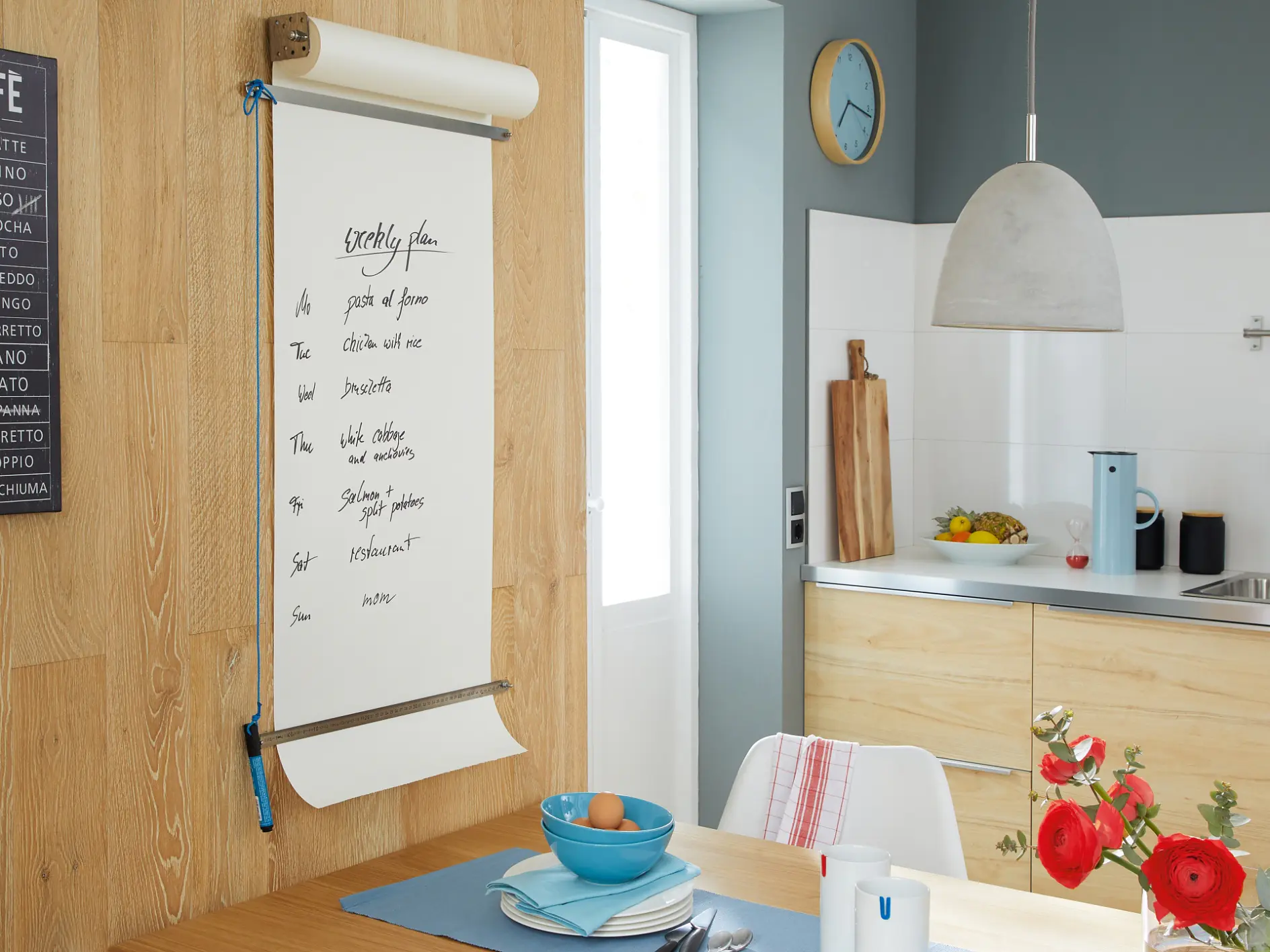 Ett modernt kök med ljusa träskåp och ett litet matbord. En stor anteckningsbok på väggen listar kaffedrycker, säkert fastsatt med tesa-tejp. På bordet finns en blå tallrik, skål och kopp. Röda blommor står i en vas och frukt ligger på bänken. En pendellampa hänger ovanför. (This text has been generated by AI)