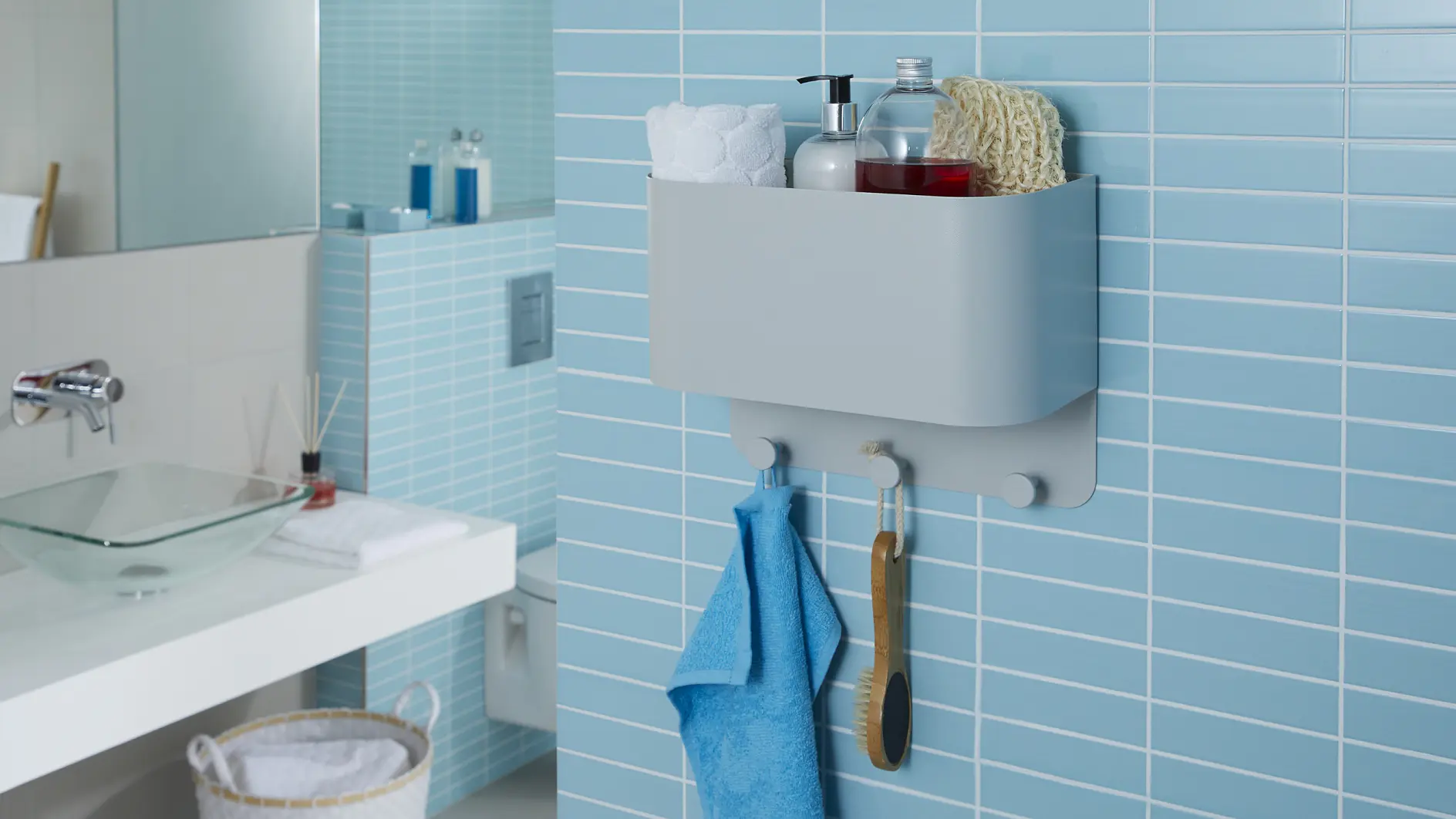 Badrum med ljusblå kaklade väggar. En väggmonterad organisatör, fäst med tesa-tejp, håller en handduk, borste, tvåldispenser och toalettartiklar. Nedanför hänger en blå handduk. En vask med glashandfat och en tvättkorg syns i bakgrunden.